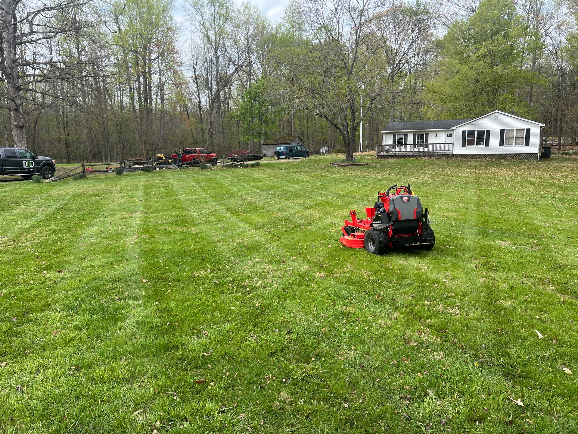 A red riding lawnmower cutting grass in a large yard next to a white house with trees in the background.