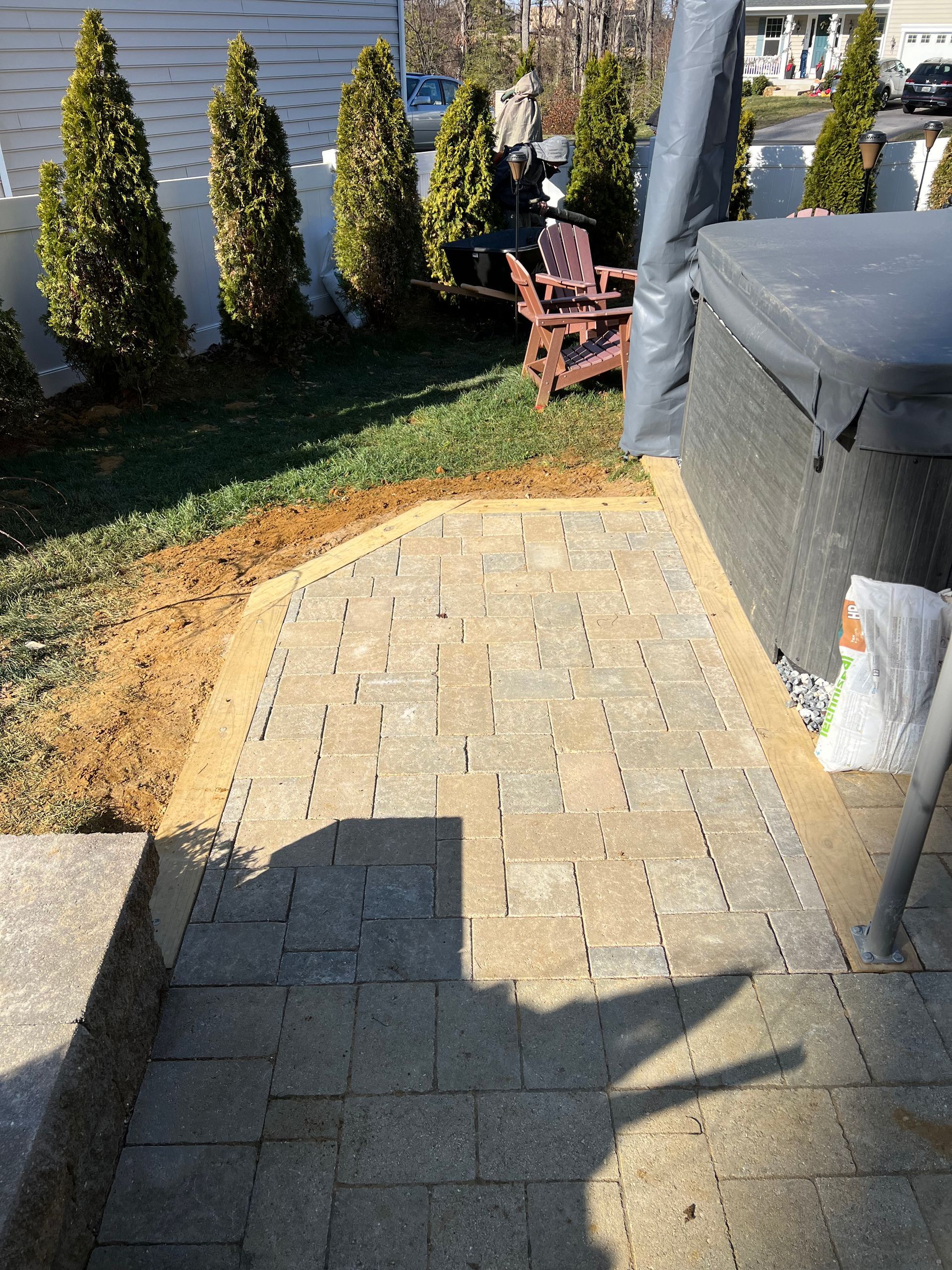 Paver patio next to a hot tub. Green grass, shrubs, and a white fence are in the background.