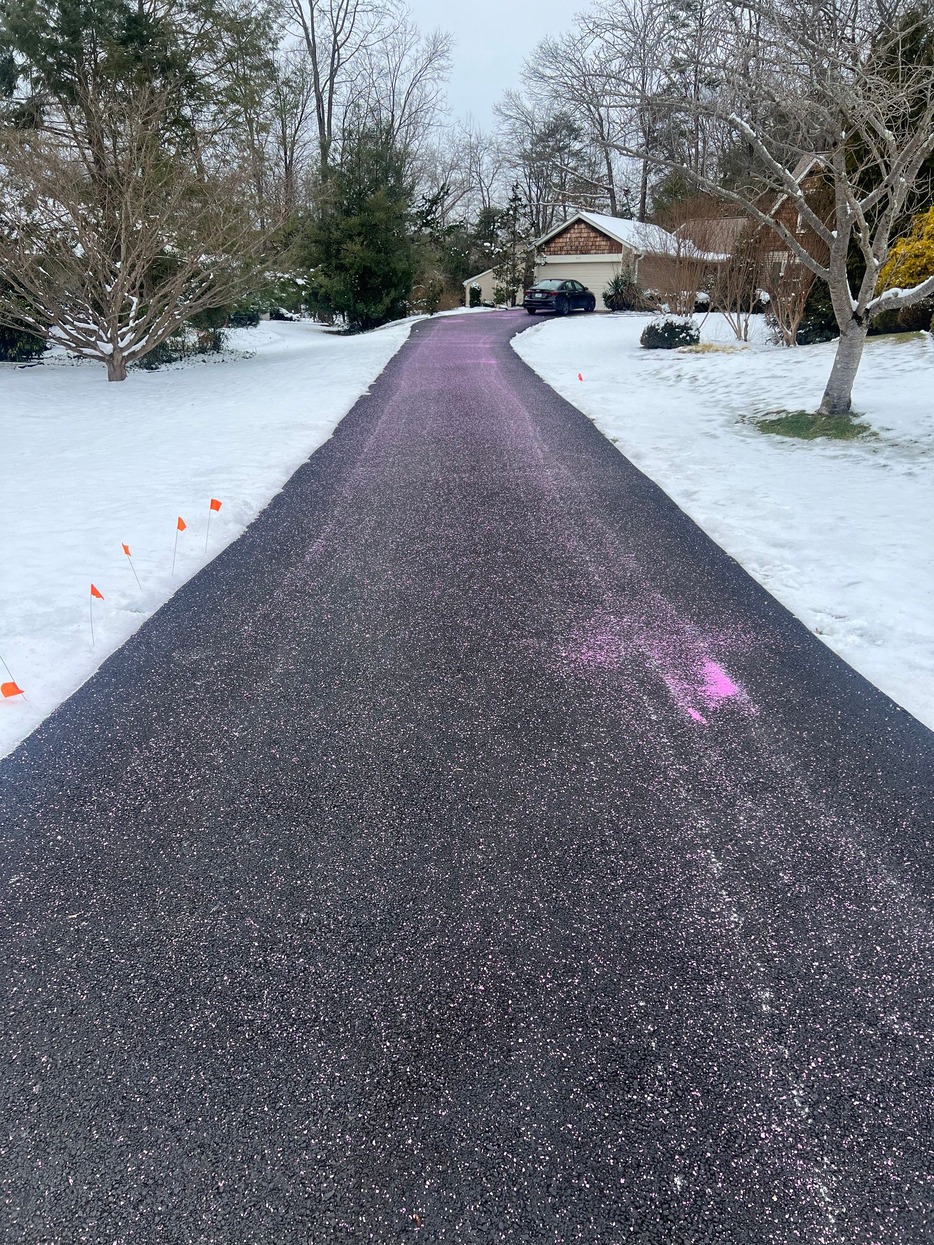 Black asphalt driveway with pink markings, flanked by snow-covered lawn. House visible in the background.