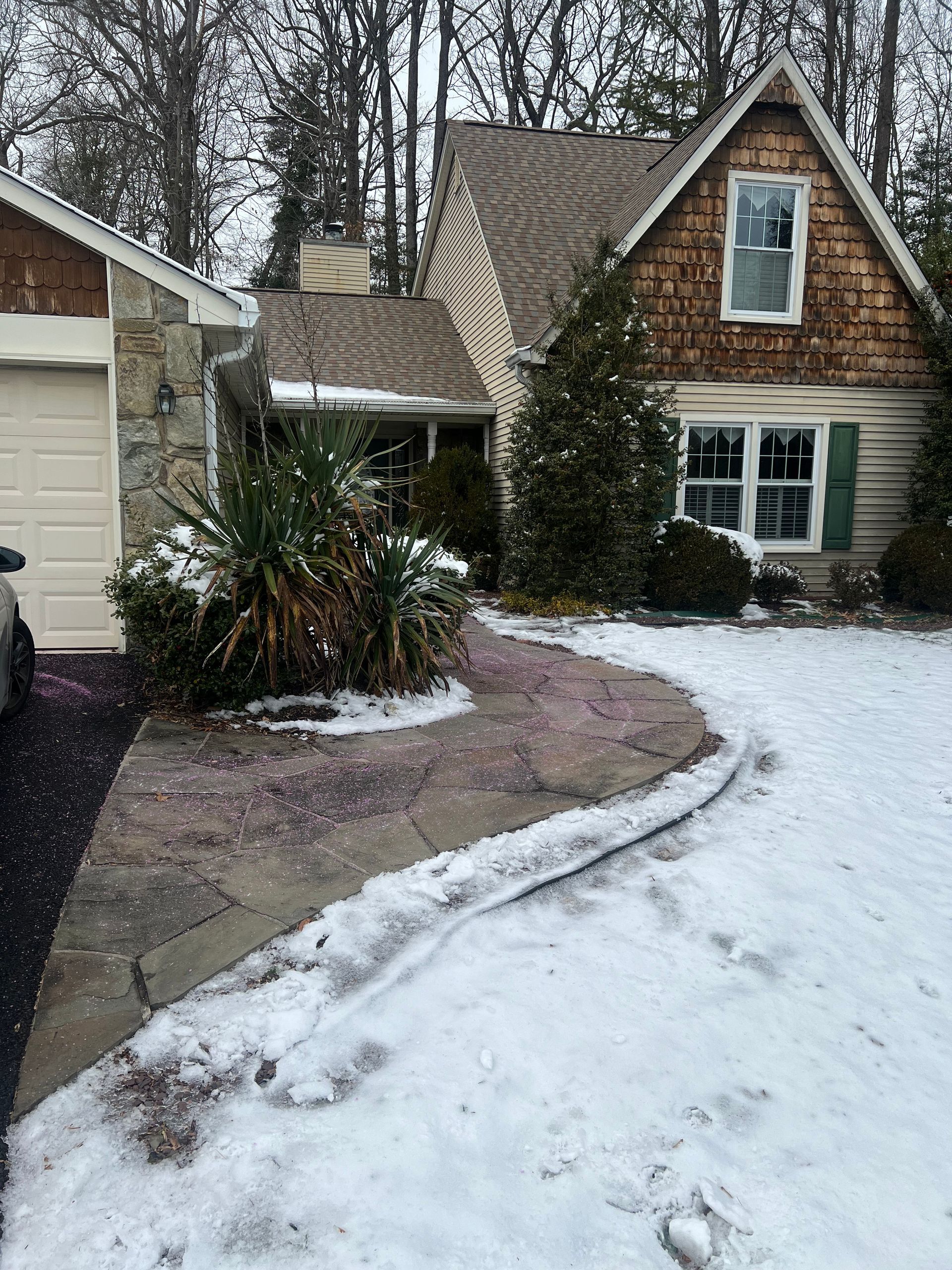 Snowy house exterior with a curved walkway. Stone garage to the left. Brown shake siding on the roof gable.