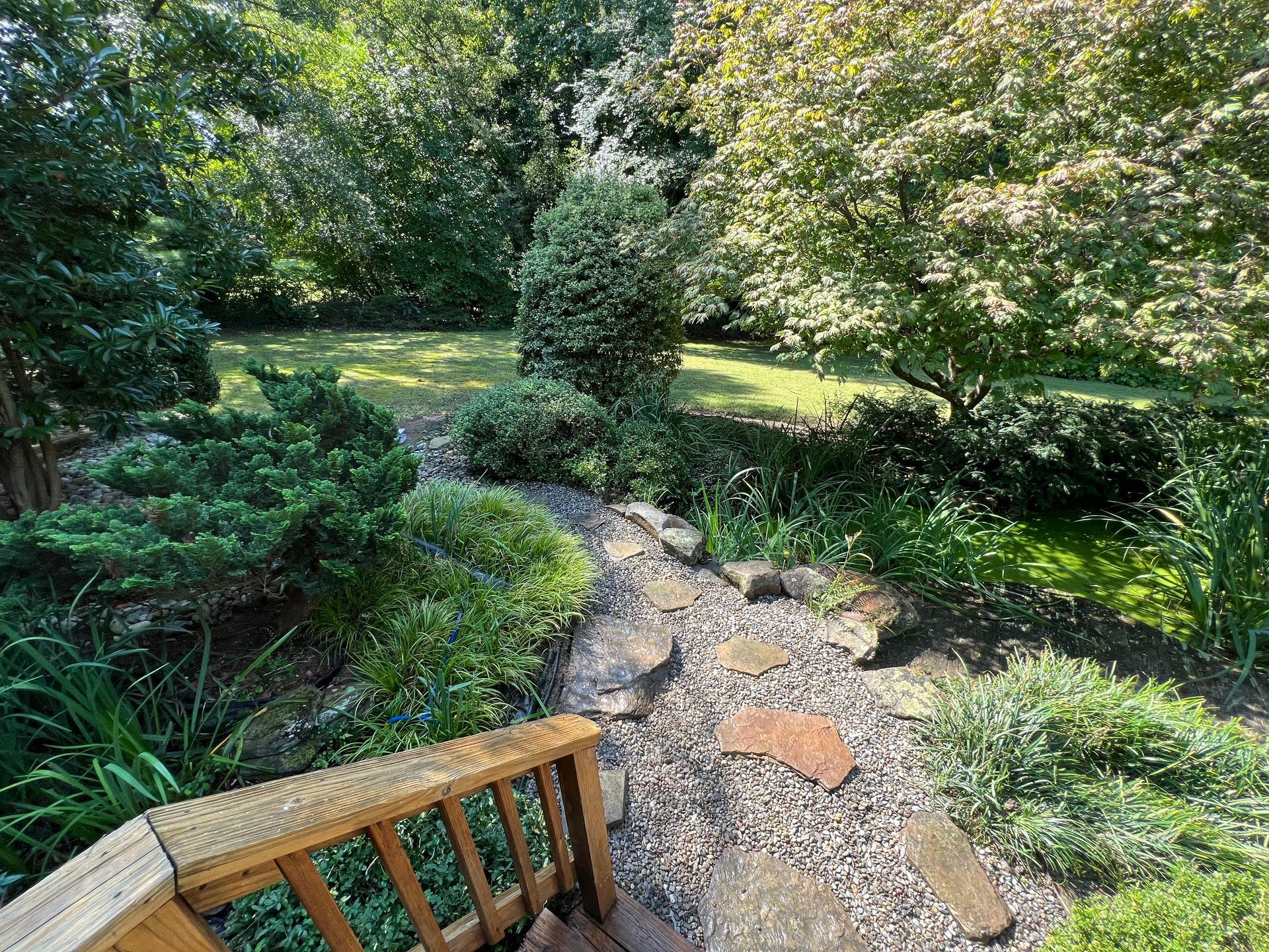 Stone path through a lush green garden, with bushes, trees, and a wooden railing in the foreground.
