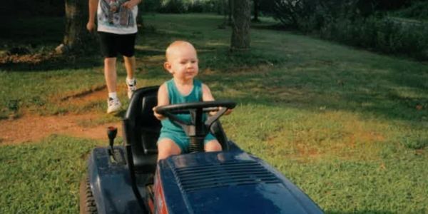 A baby sits on a lawnmower, holding the steering wheel. A person walks behind it on the grass.