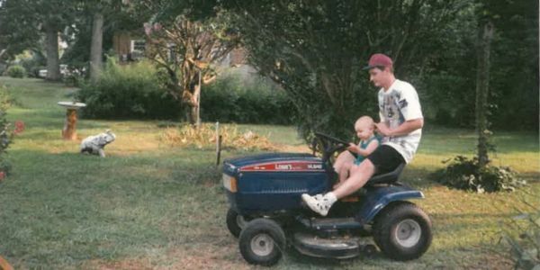 Man and baby ride on a blue lawn tractor in a grassy yard.