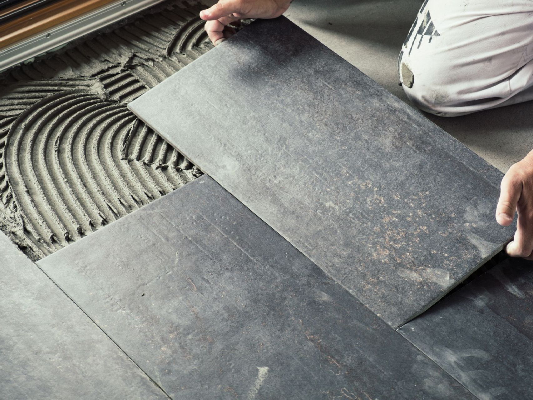 Hands installing large gray floor tiles over fresh mortar in a room.