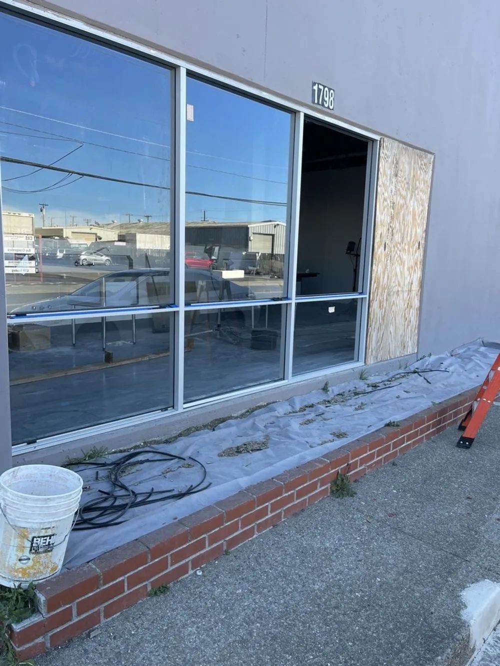 Exterior view of building with large windows. One window frame is open; brick ledge and bucket in front.