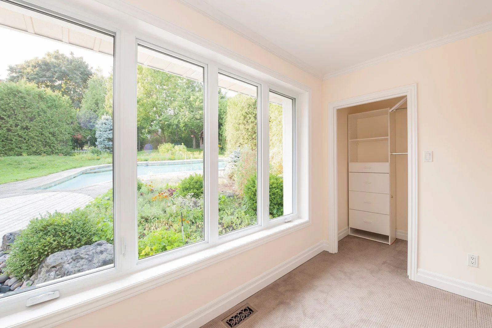 Room with large windows overlooking a backyard with a pool. White trim, beige walls, and carpet. Open closet.