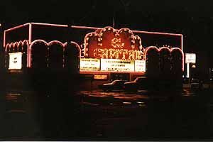 A movie theater is lit up at night with cars parked in front of it.
