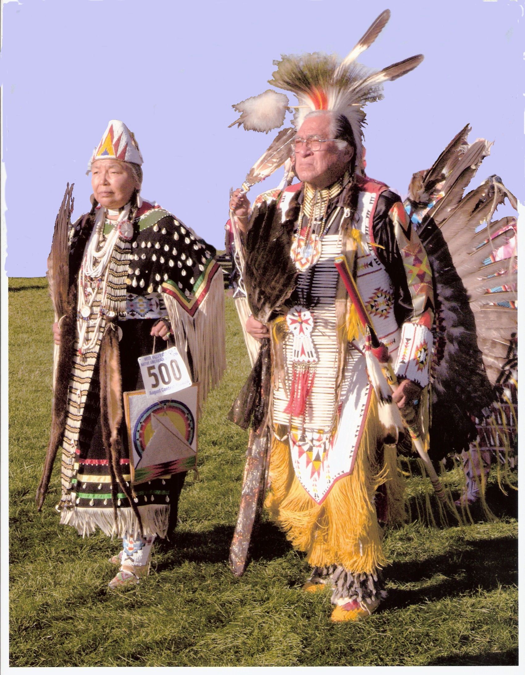 Two men in native american costumes are walking in a grassy field.