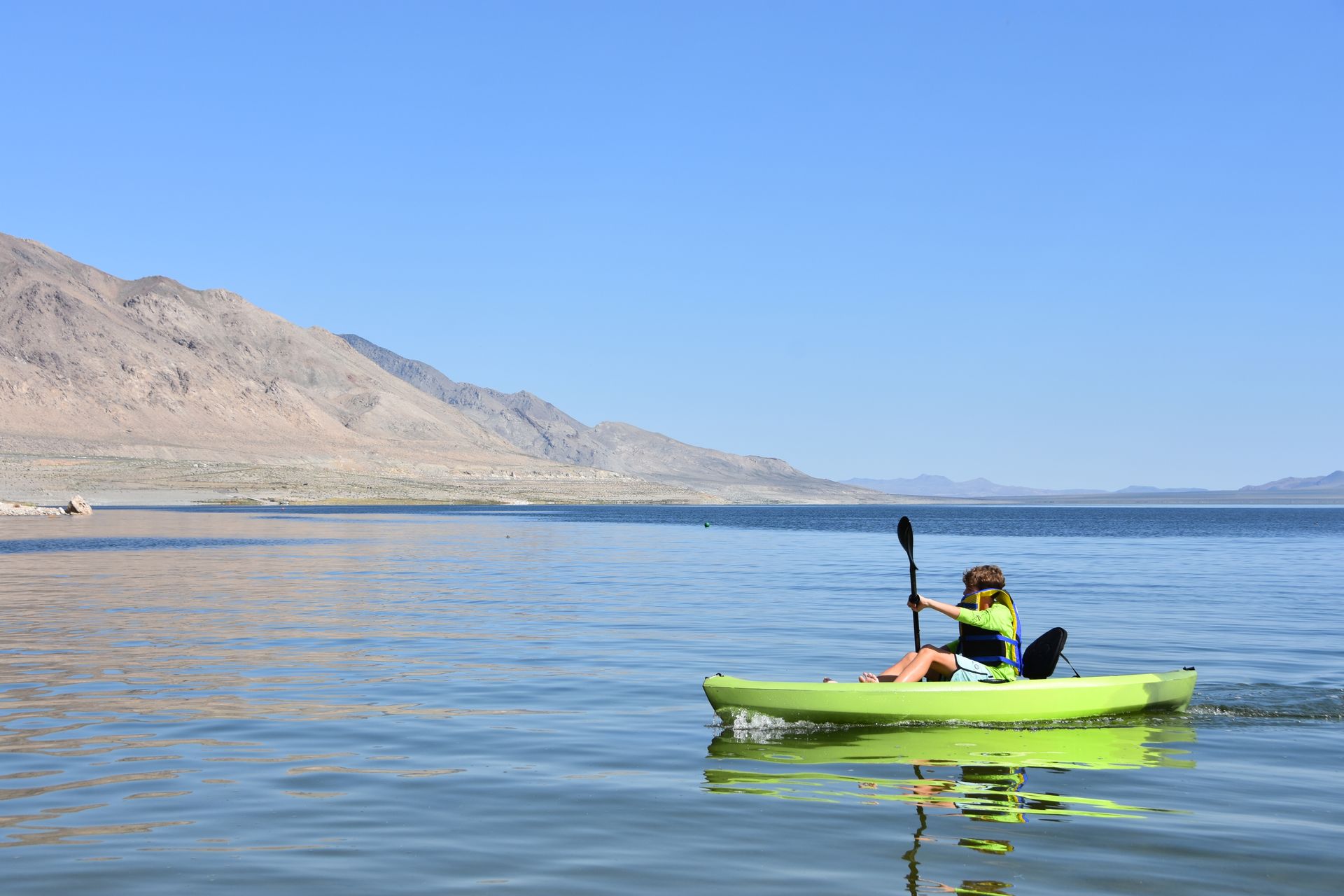 A person is paddling a kayak on a lake with mountains in the background.