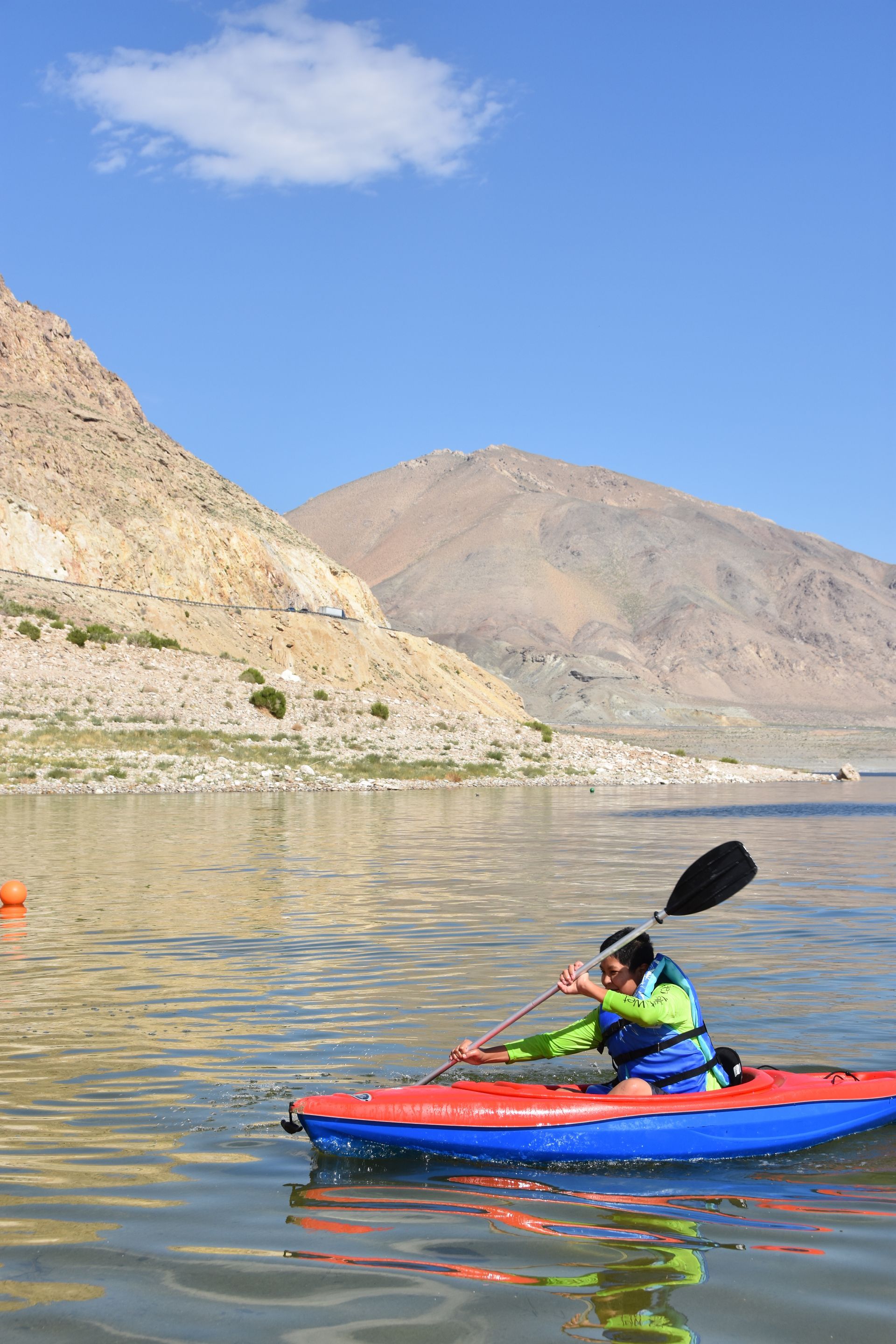 A person is paddling a kayak on a lake with mountains in the background.