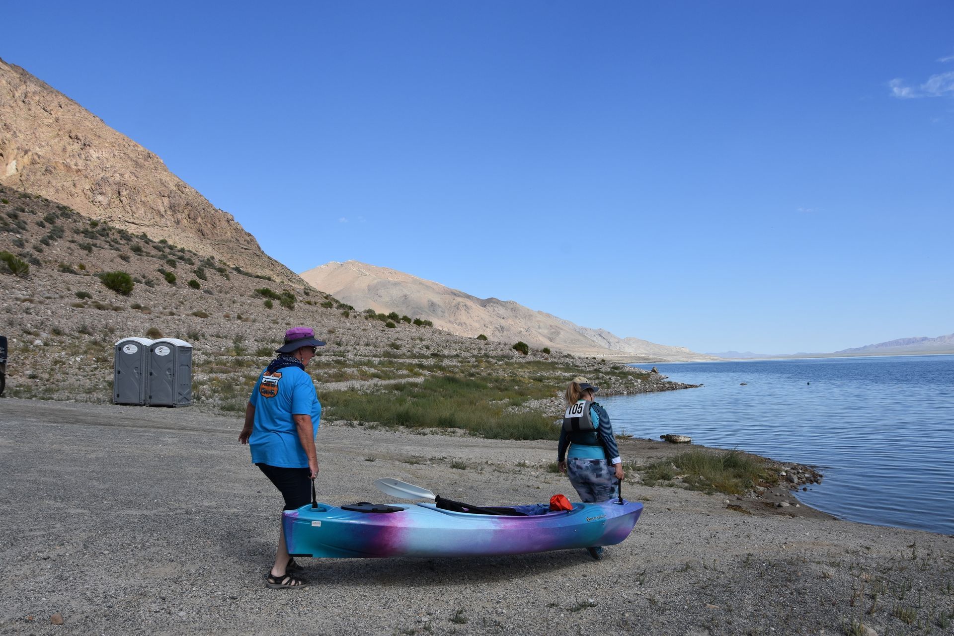 Two people are pushing a kayak on the beach near a lake.