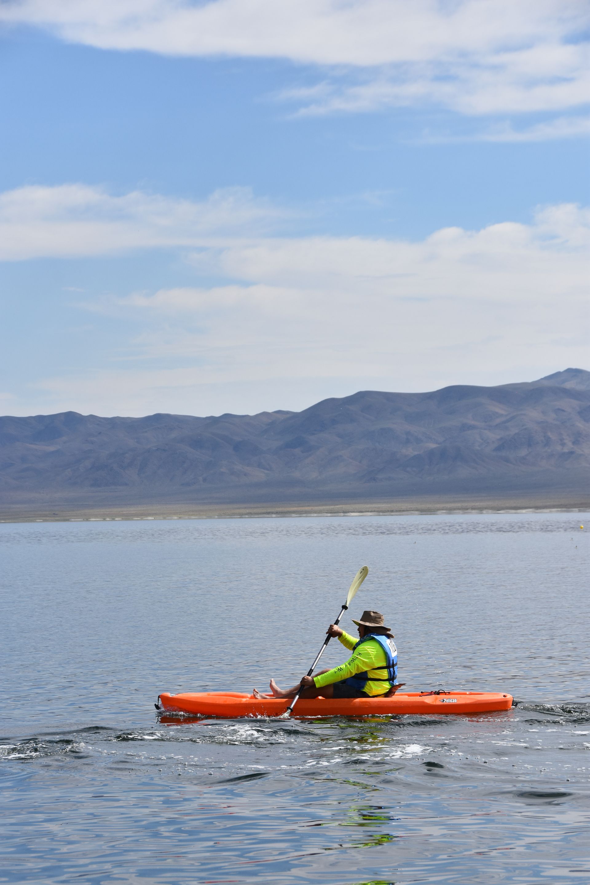 A person is paddling a kayak on a lake with mountains in the background.