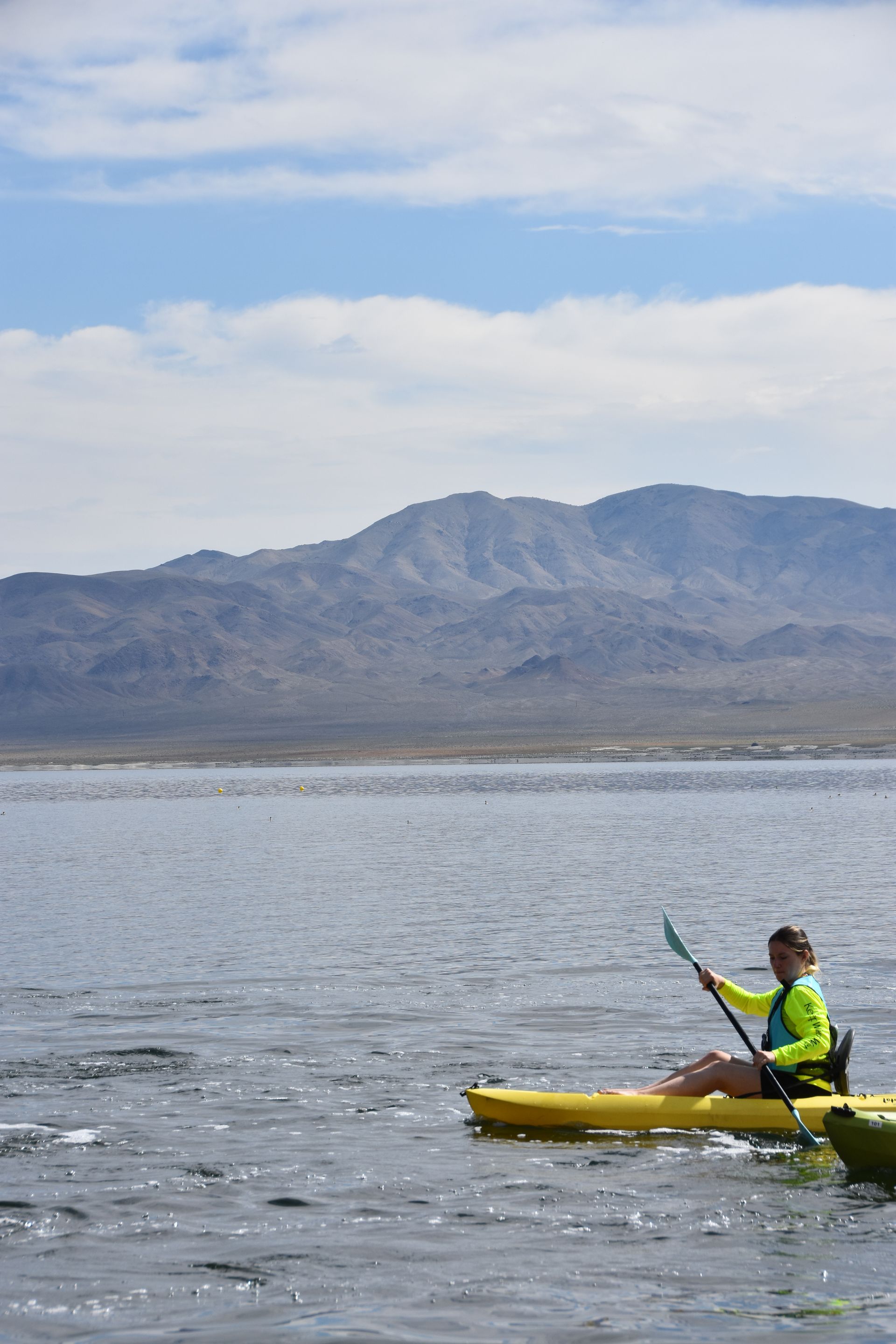 A person is paddling a kayak on a lake with mountains in the background.