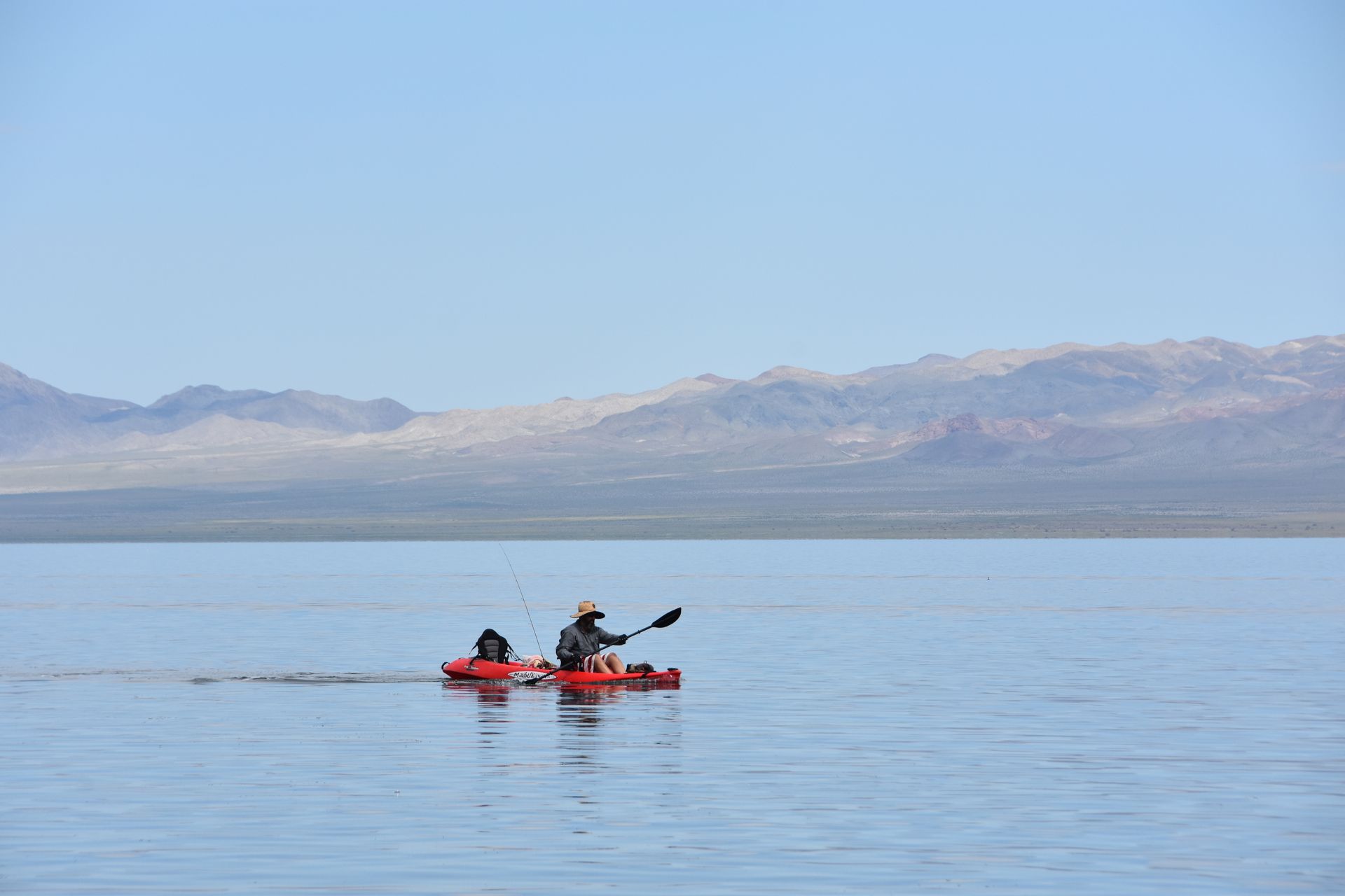 A man is paddling a red kayak on a lake with mountains in the background.