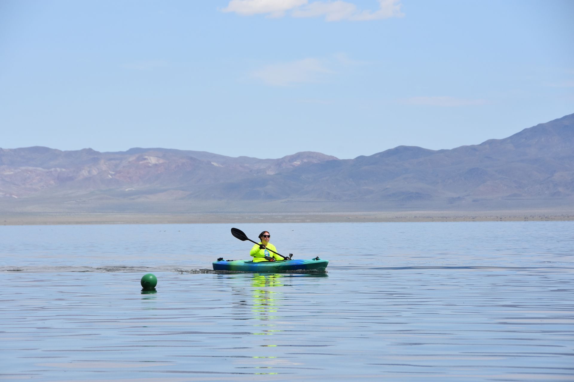 A person is paddling a kayak on a lake with mountains in the background.