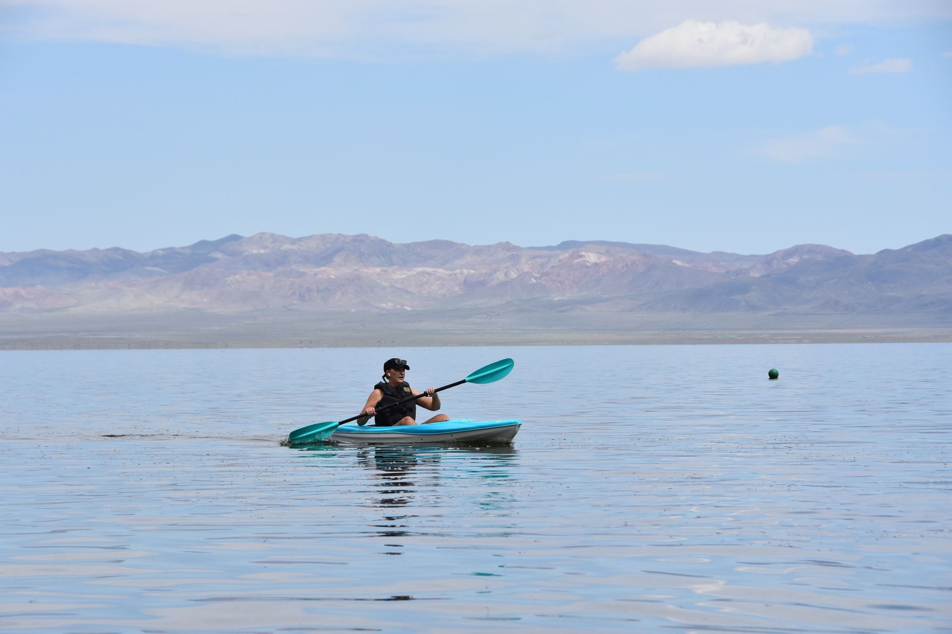 A man is paddling a kayak on a lake with mountains in the background.