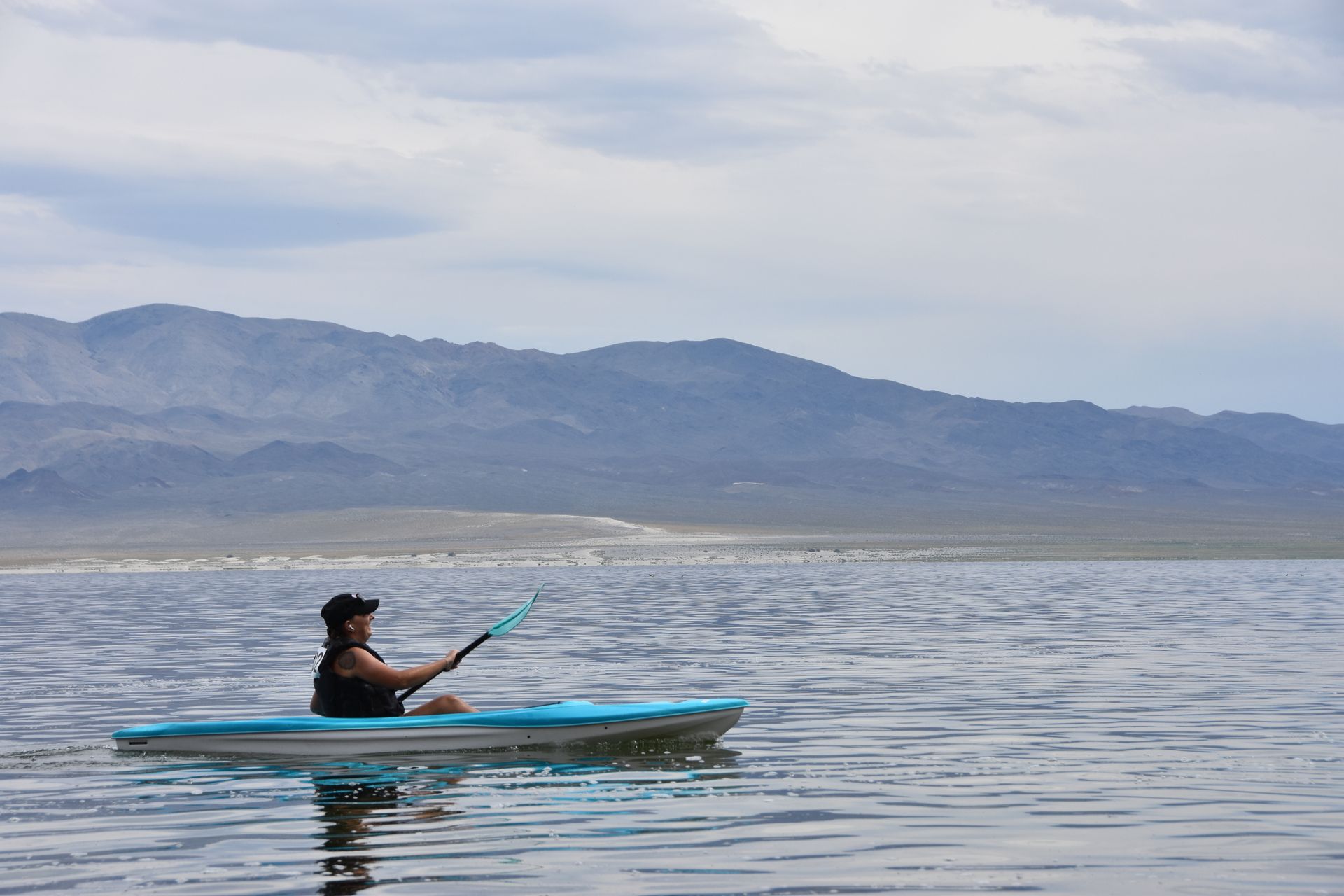 A man is paddling a kayak on a lake with mountains in the background.