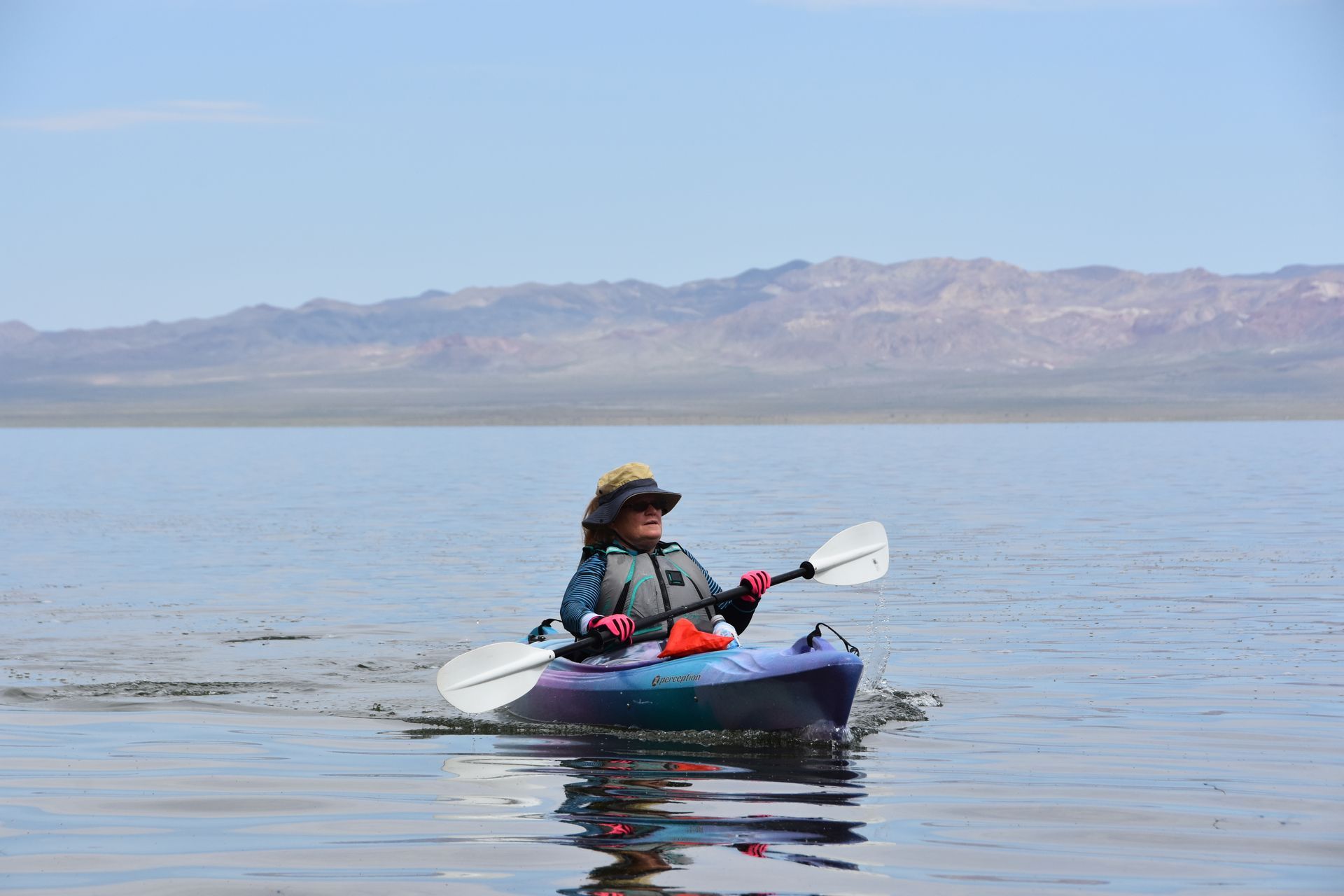 A person is paddling a kayak on a lake with mountains in the background.