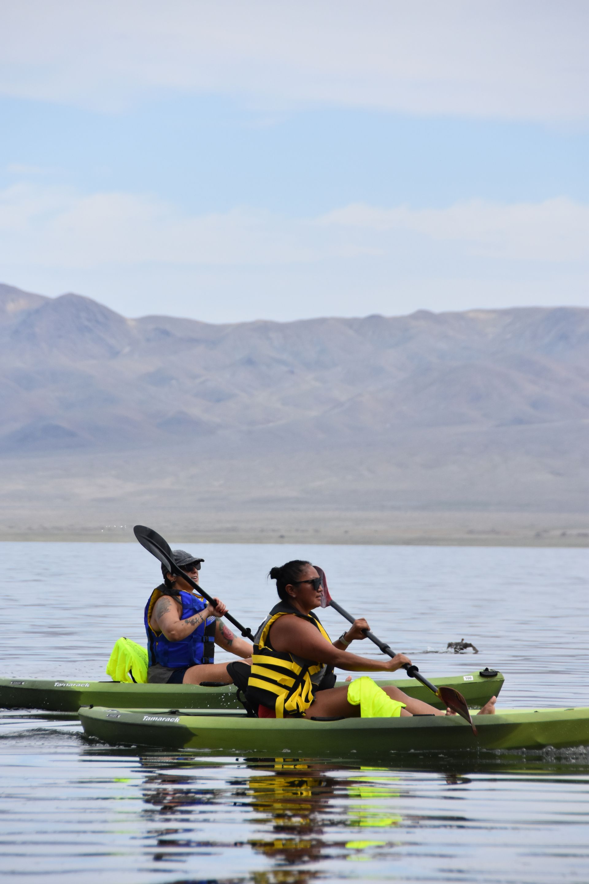 Two people are paddling kayaks on a lake with mountains in the background.