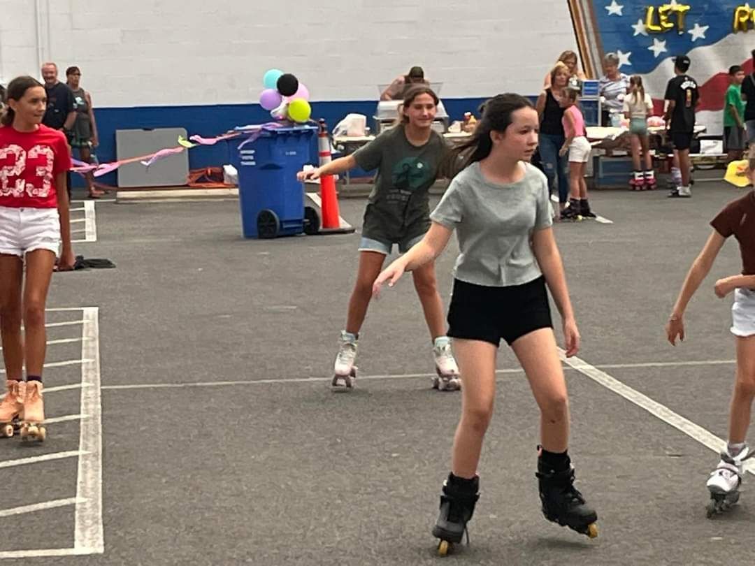 Girls roller skating, wearing athletic clothes, at Hawthorne's Mainstreet Skate Night 2024, with decorations and a crowd.
