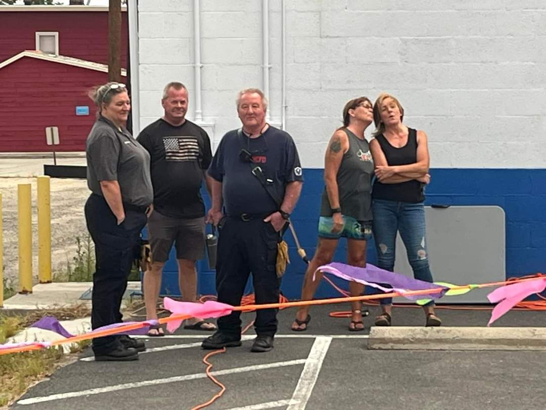 A group of people watching skaters during Skate Night, including a firefighter, by a building with pink and purple ribbons.