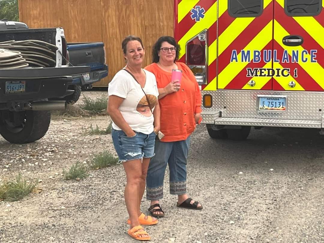 Two women standing by an ambulance and a truck. One wears jean shorts and a tee, the other an orange shirt and jeans. 