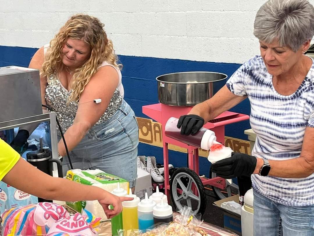 Two women selling treats during Hawthorne's Mainstreet Skate Night 2024. One adds syrup to a treat, another points to a cake.
