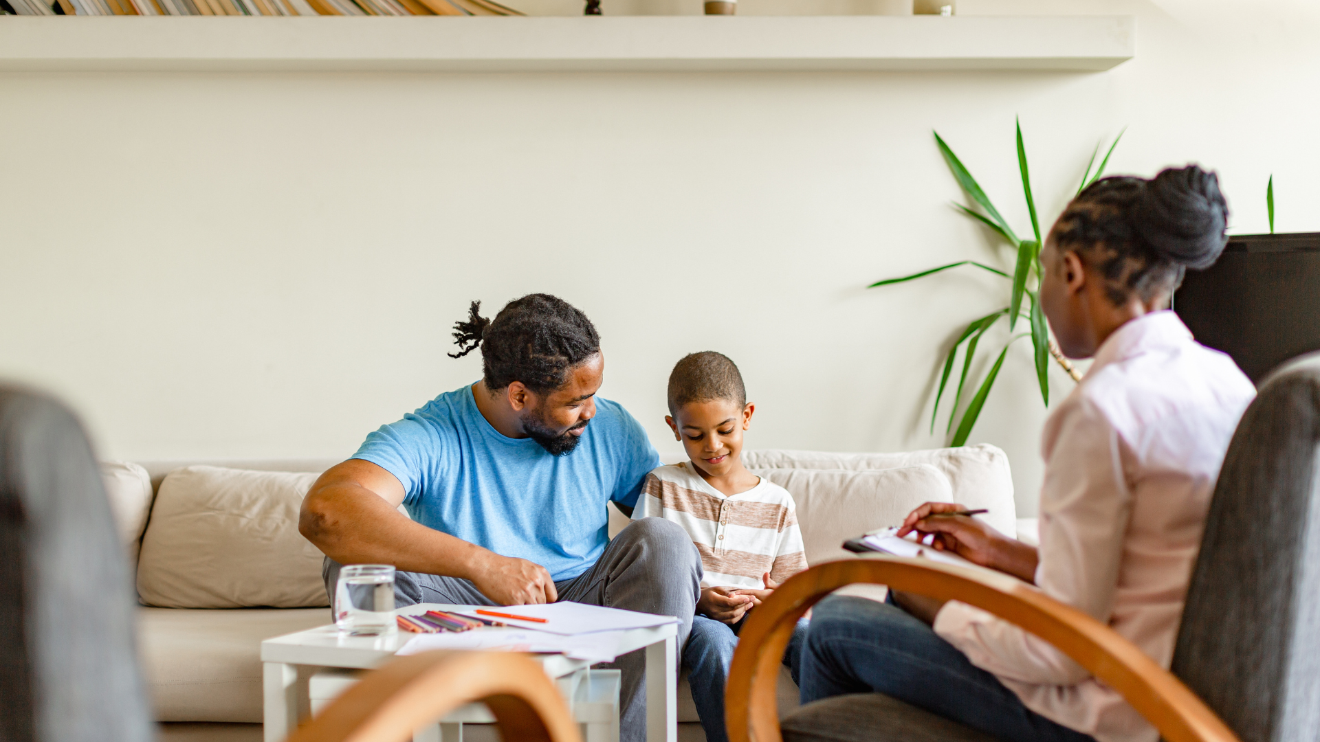 A man is sitting on a couch talking to a woman who is writing on a clipboard.