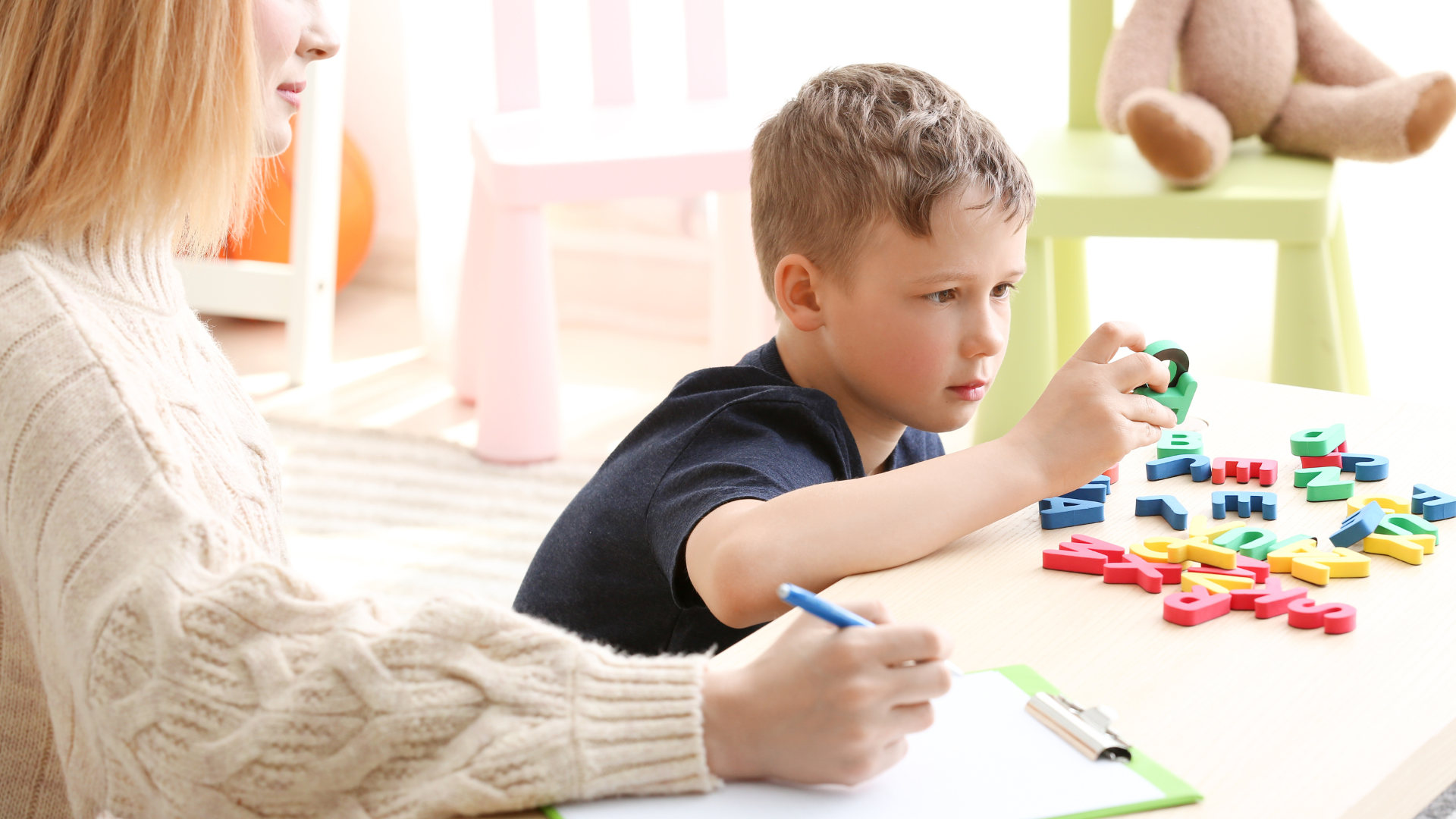 A child plays with colorful toys while a therapist takes notes at a table in a bright room.