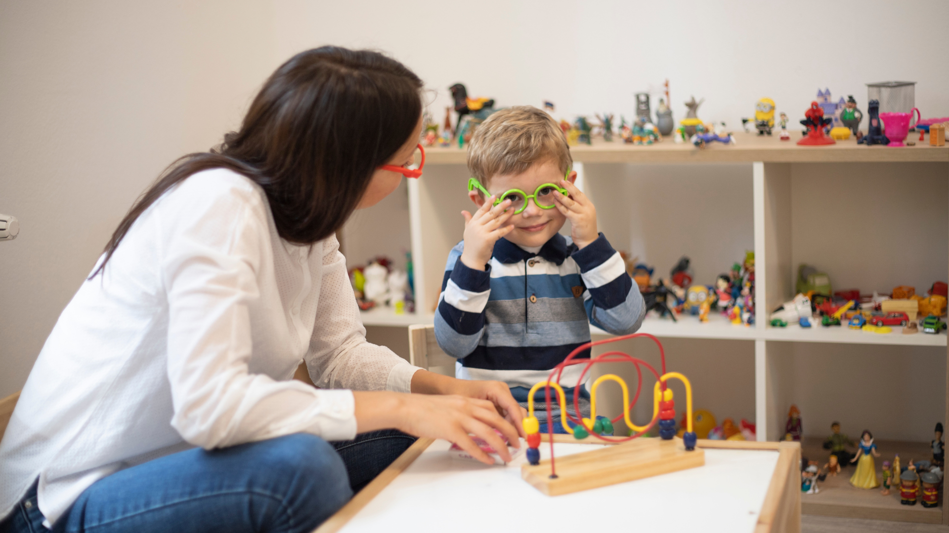 Woman and child in playroom; child wearing green glasses, playing with a toy maze.