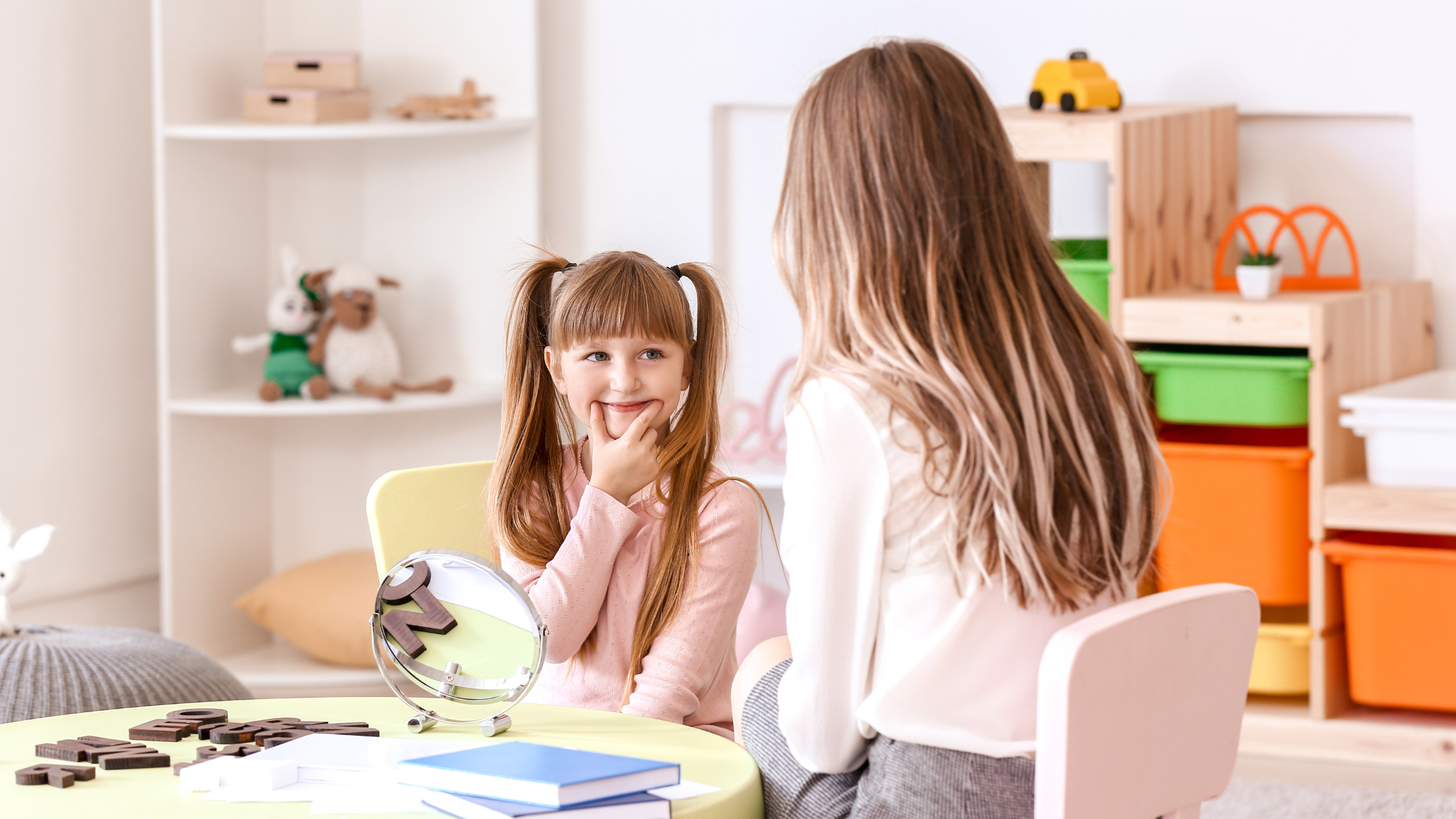 Girl with pigtails smiles at a woman in a therapy session. Bright room with toys, table, and chairs.