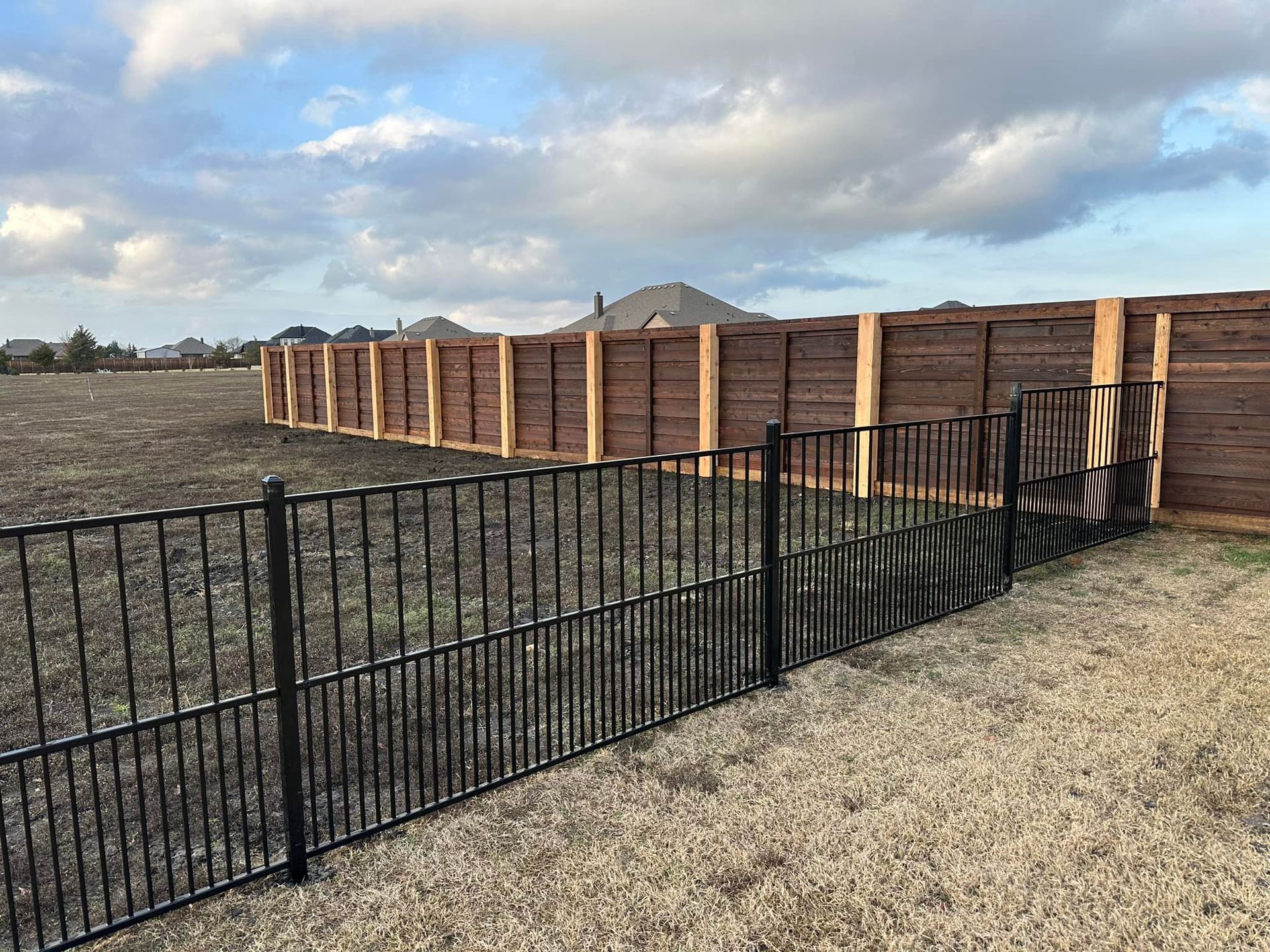 Black metal fence in front of a tall wooden fence, in an open field, under a cloudy sky.