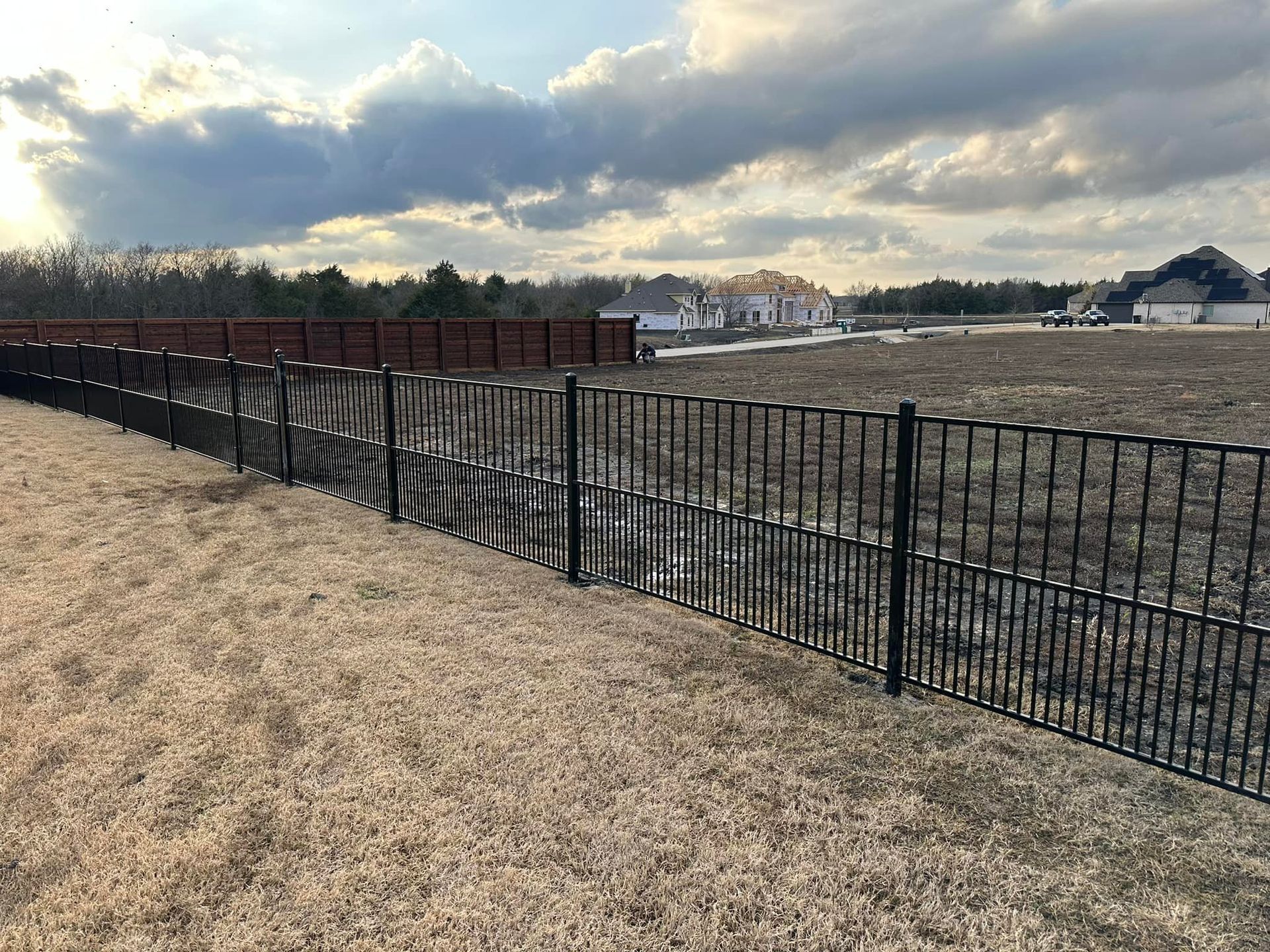 Black metal fence along dry grass, with a brown fence and houses in the distance under a cloudy sky.