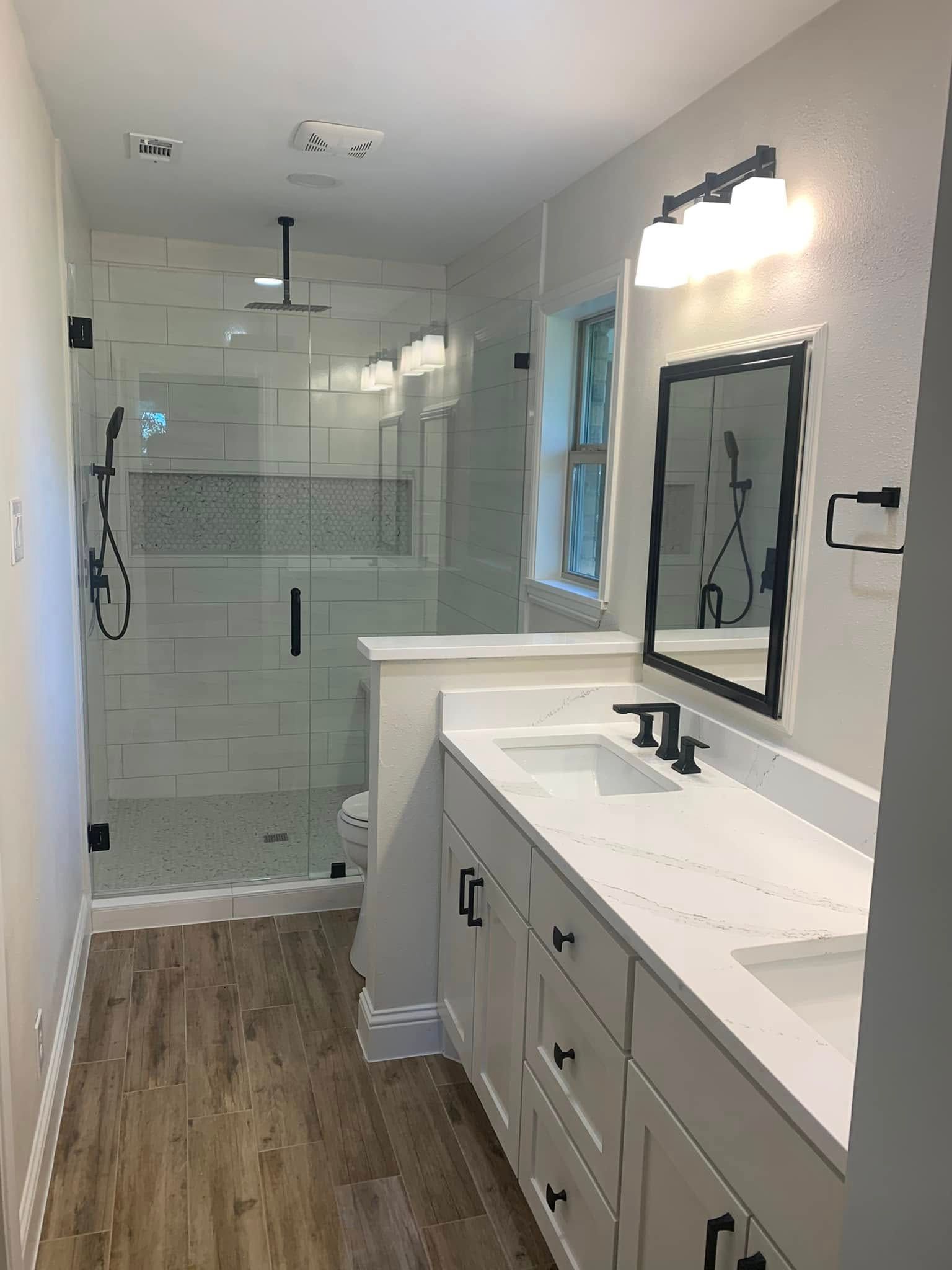 Modern bathroom with glass shower, double vanity, and light wood flooring. White and black accents.