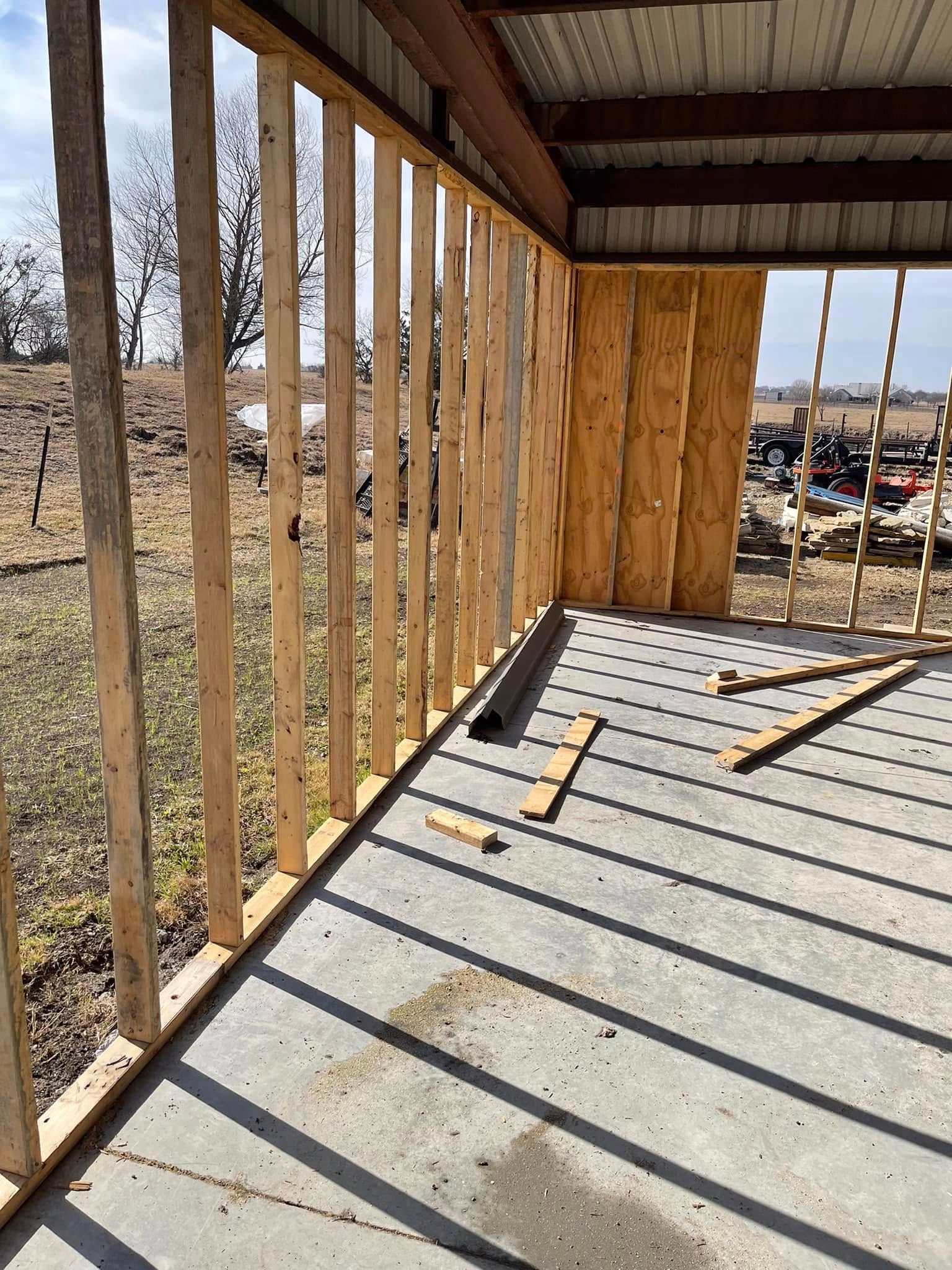 Construction of wooden wall studs on concrete floor, metal roof visible, outdoors.