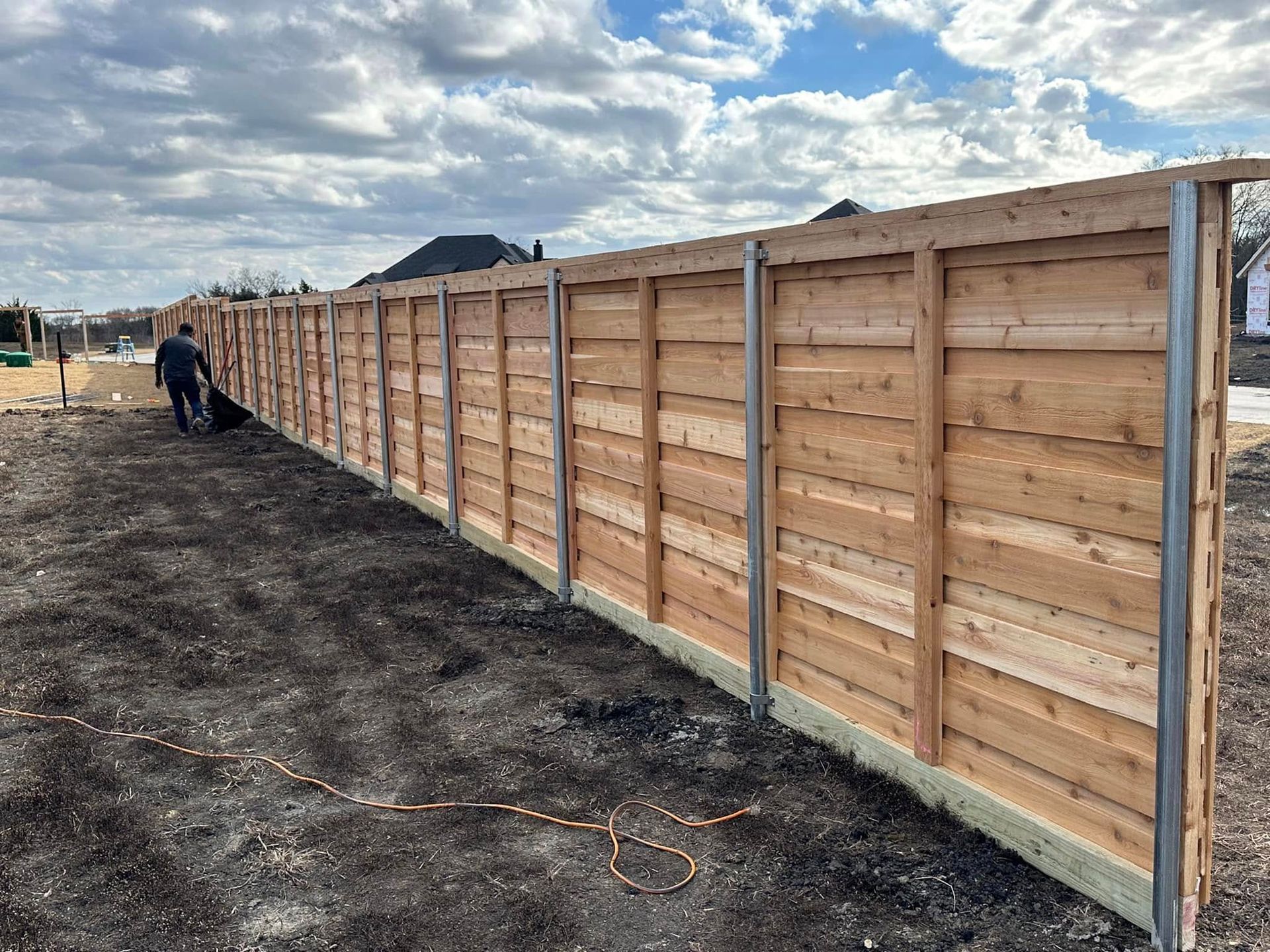Long wooden privacy fence with steel posts; person in dark clothing walks along the side, cloudy sky above.