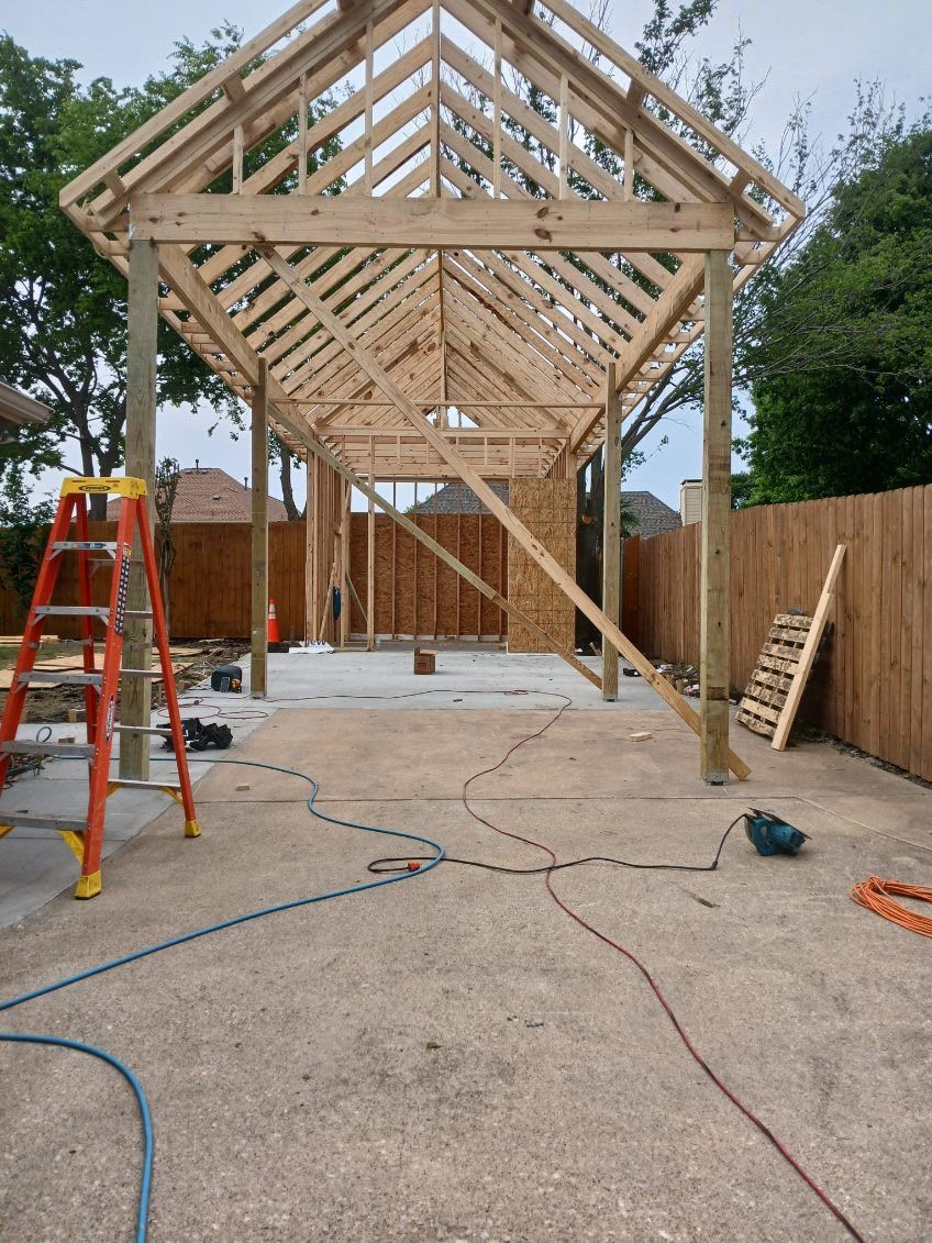 Wooden structure under construction, with visible rafters, posts, and concrete floor. Tools and ladder present.