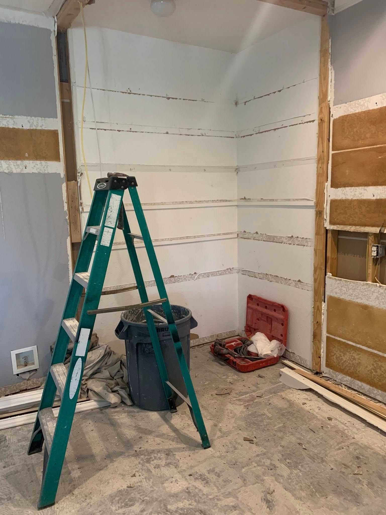 Interior room under construction, with a green ladder, trash can, and toolbox.  White-painted alcove for shelves is visible.