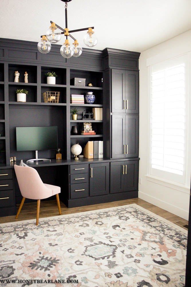 Black built-in desk and shelving in home office, with gold accents and pink chair. Includes a rug, light fixture, and window.