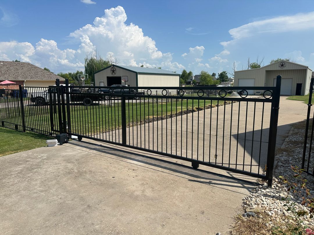 A black metal fence with a mesh screen borders a dry, brown field with houses under a cloudy sky.