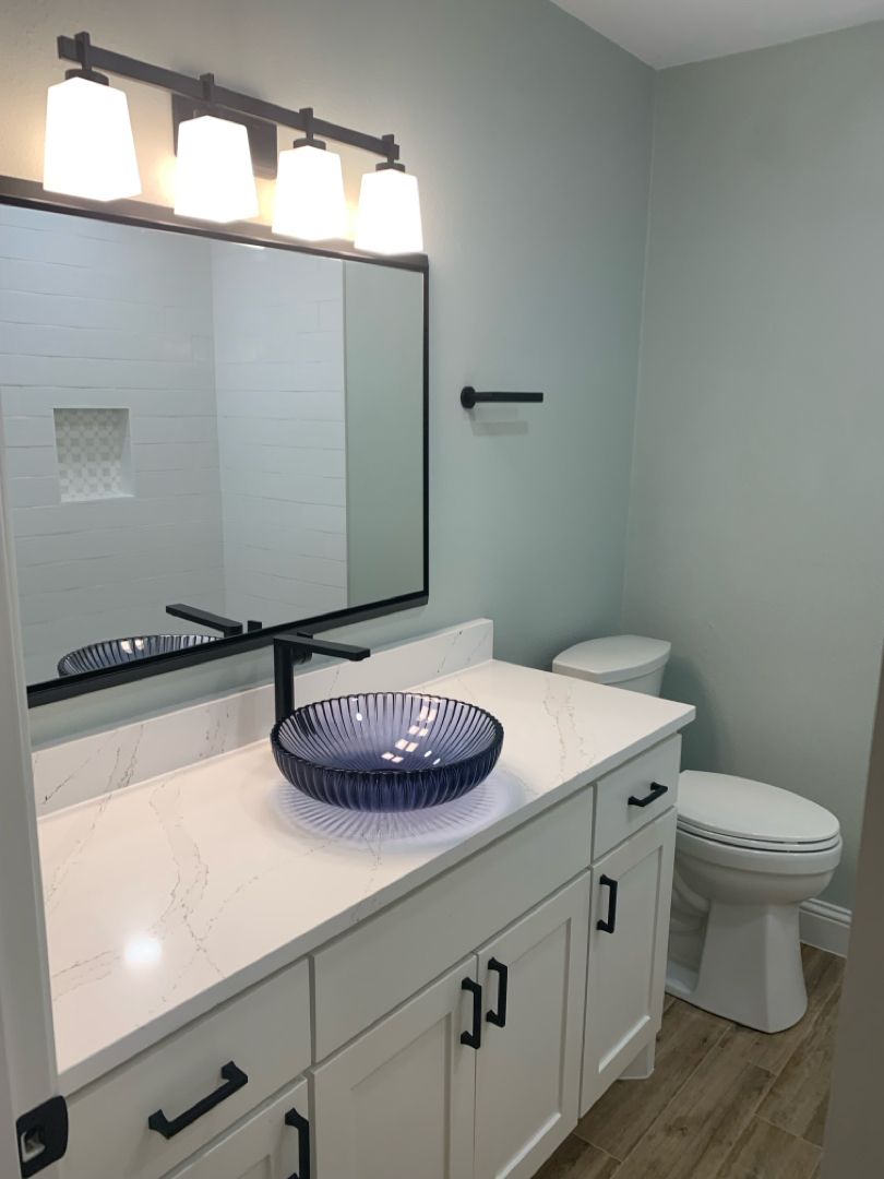 A modern bathroom featuring a white vanity with black fixtures, a mirror, and a glass-enclosed shower with wood floors.