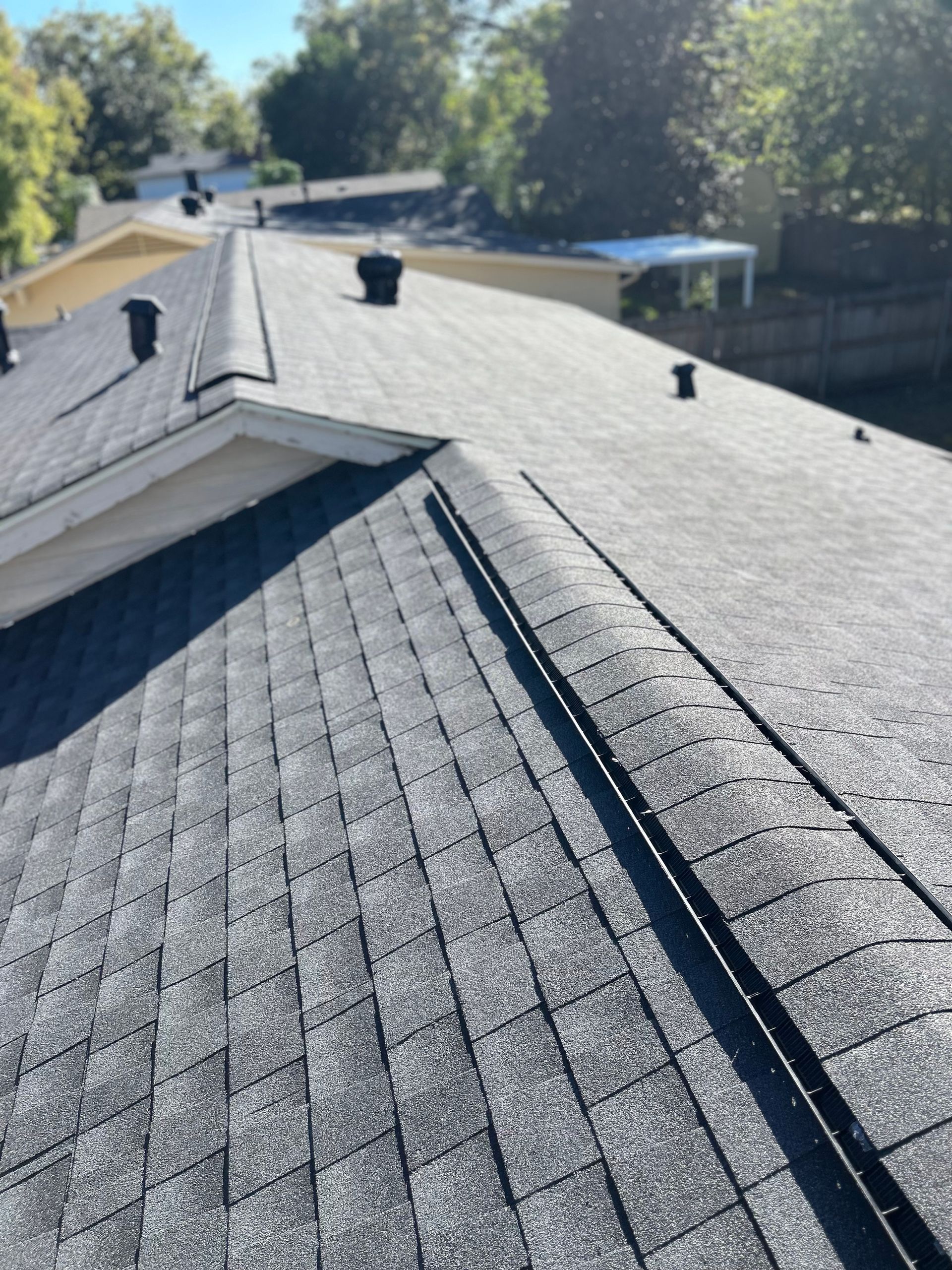 Two people installing dark asphalt shingles on the roof of a house under a clear blue sky.