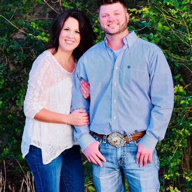 Couple smiling, posing outside. Woman wears white lace top, jeans; man wears blue shirt, jeans, belt buckle.