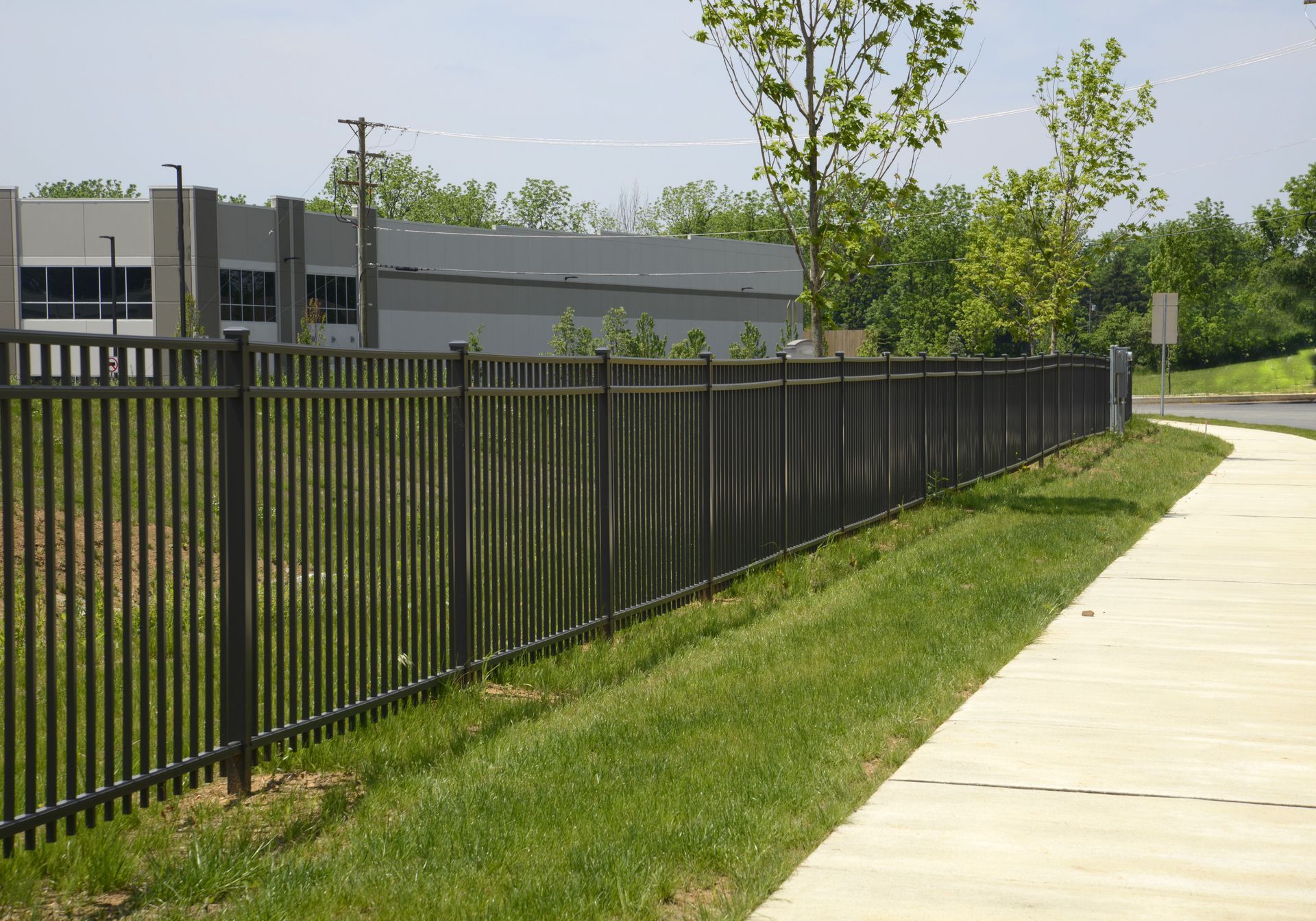 Black metal fence bordering a sidewalk and grassy hill, with a building and trees in the background.