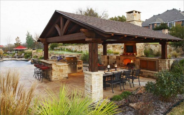 Outdoor kitchen and dining area with a fireplace under a wooden roof.