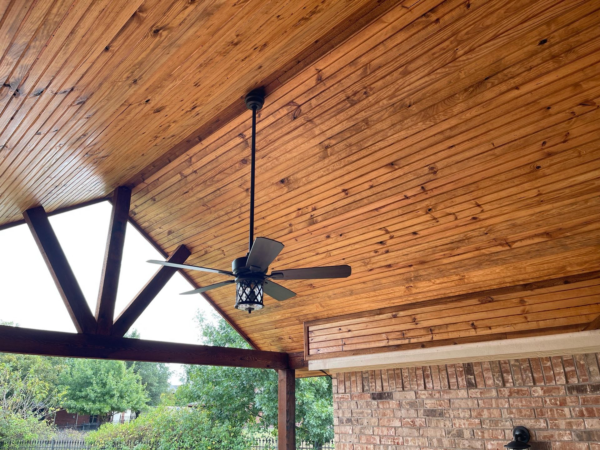 Wooden outdoor ceiling with a hanging fan, brick wall on the right, and trees visible.