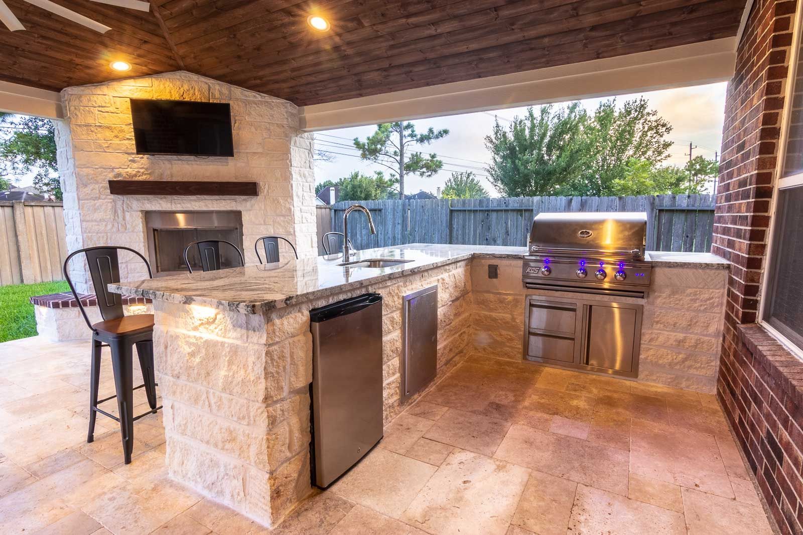 Outdoor kitchen with stone counters, grill, sink, and bar seating, under a covered patio.
