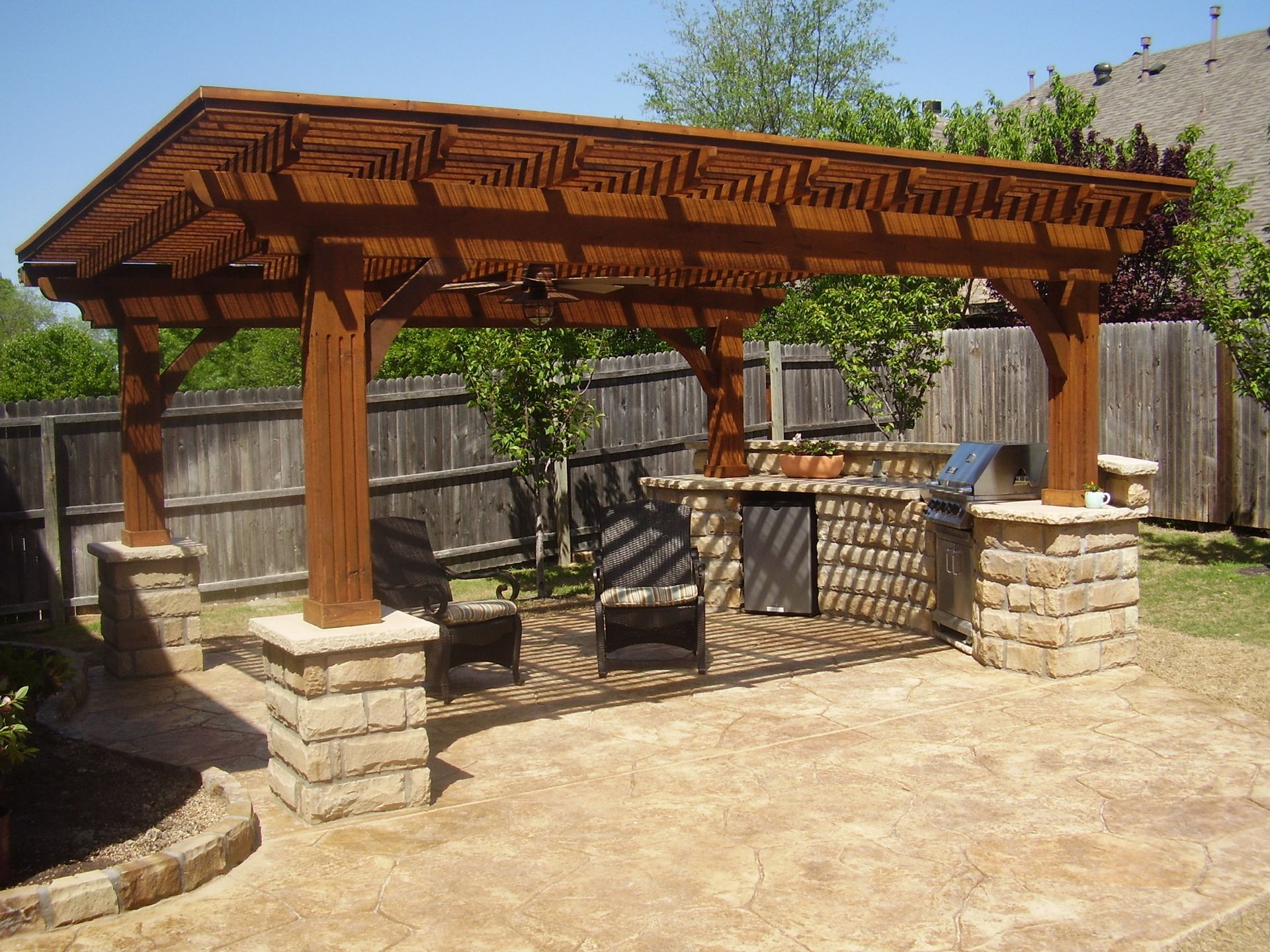 Wooden pergola over outdoor kitchen on a patio. Brown structure with grill, seating, and stone pillars.