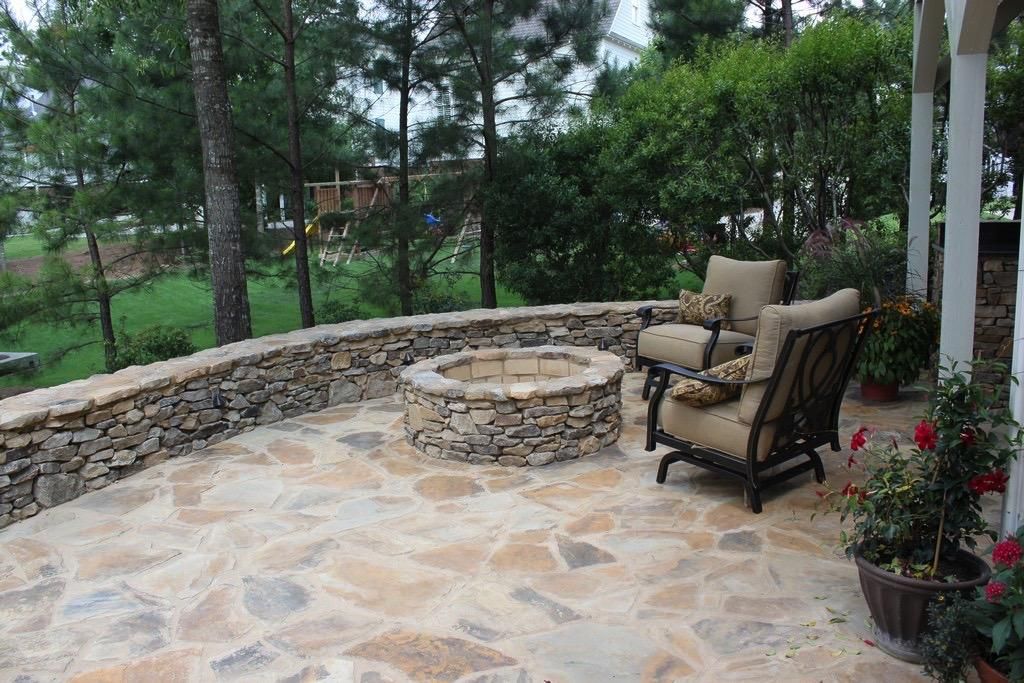 Stone patio with fire pit, two chairs, and a low stone wall, surrounded by trees.