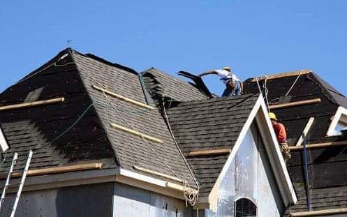 Roofers working on a house roof under a blue sky, installing dark asphalt shingles.
